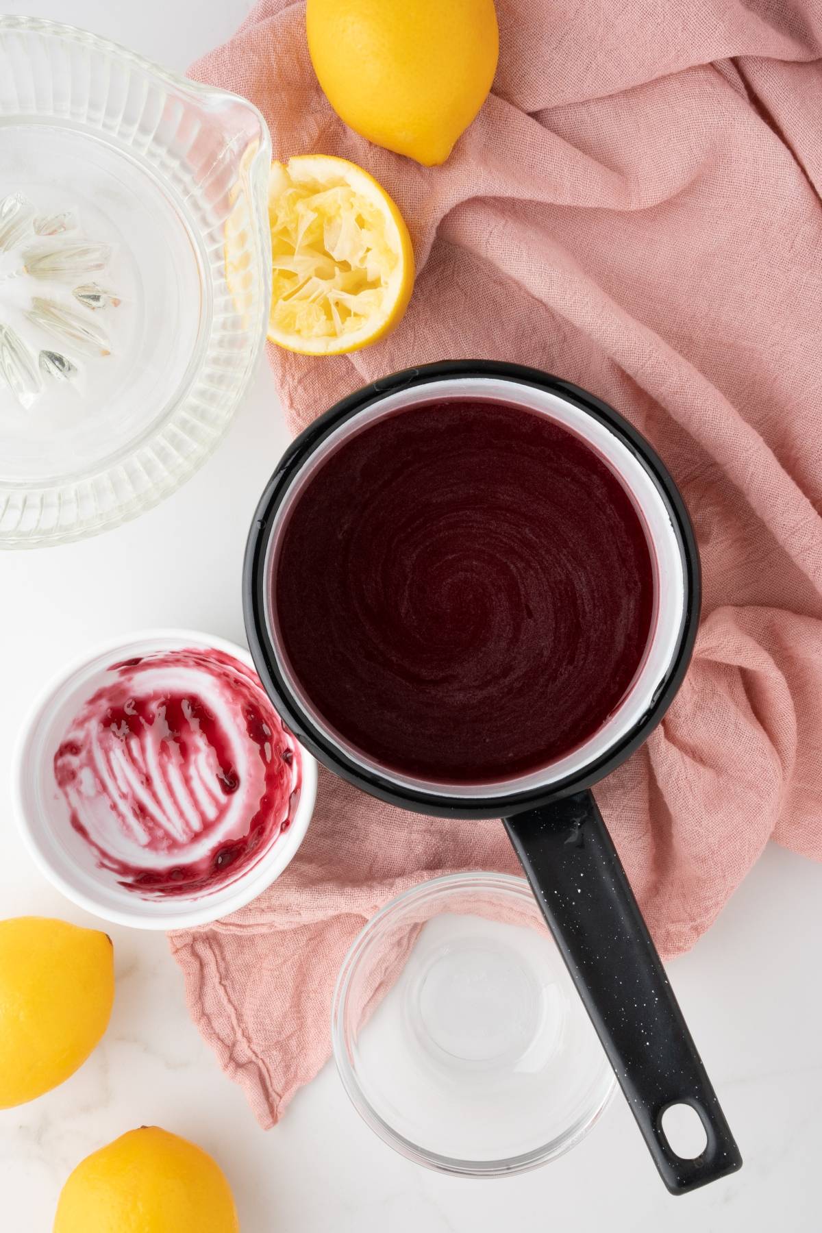 Small pot of berry sauce, empty dish, squeezed lemons, juicer, and pink cloth on a white surface—perfect for crafting a festive Lemon Berry Prosecco Cocktail or adding flavor to your New Year’s Eve cocktail.