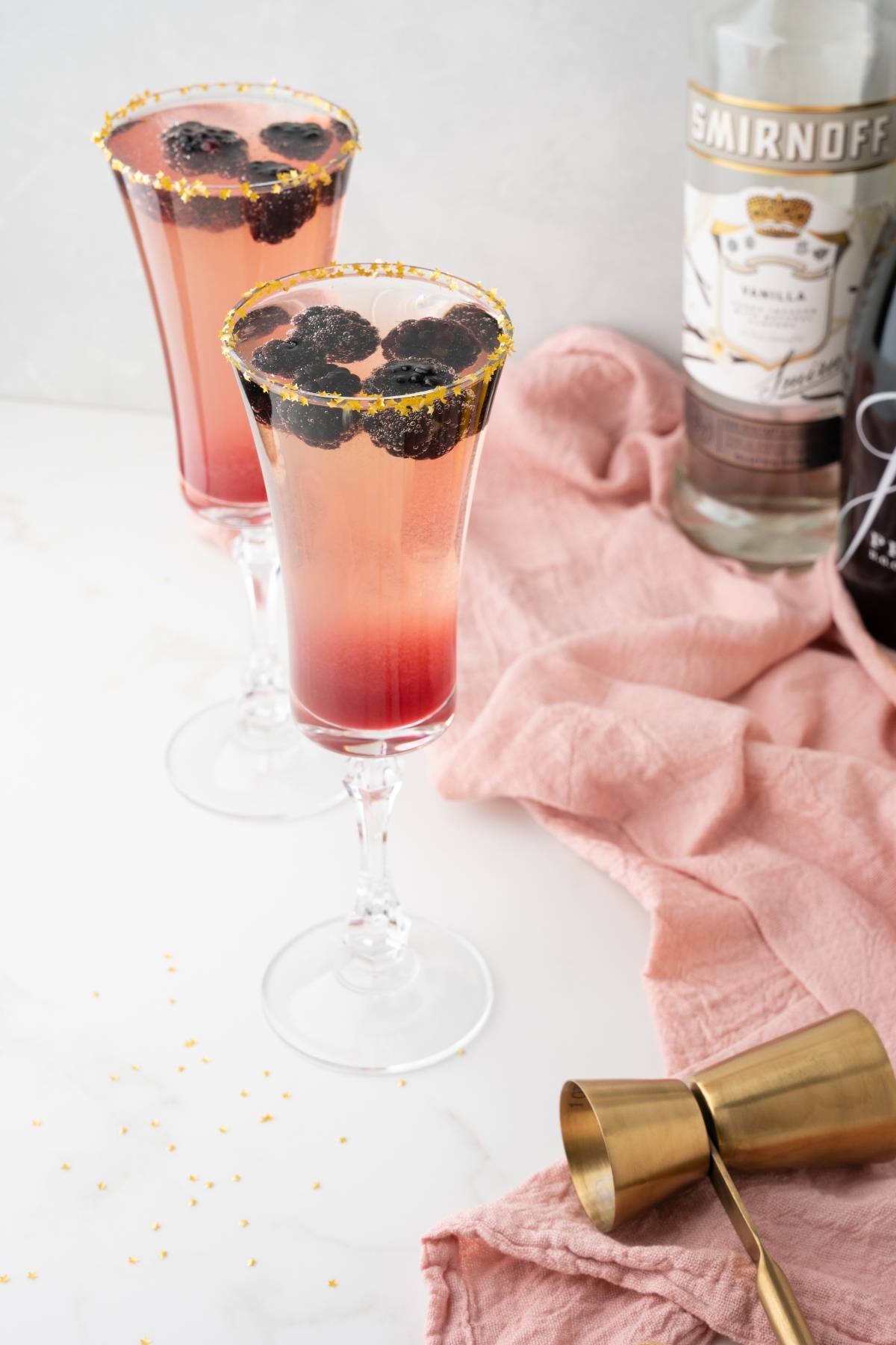 Two berry cocktails in fluted glasses with gold rims and blackberries, featuring a bottle of Smirnoff in the background—a festive Lemon Berry Prosecco Cocktail perfect for your next New Year’s Eve cocktail celebration.