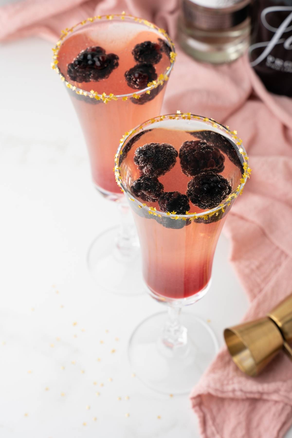 Two glasses of a simple prosecco drink with pink sparkle, blackberries, and sugared rims sit on a light surface with a pink cloth—perfect for a festive Christmas or New Year’s Eve cocktail.