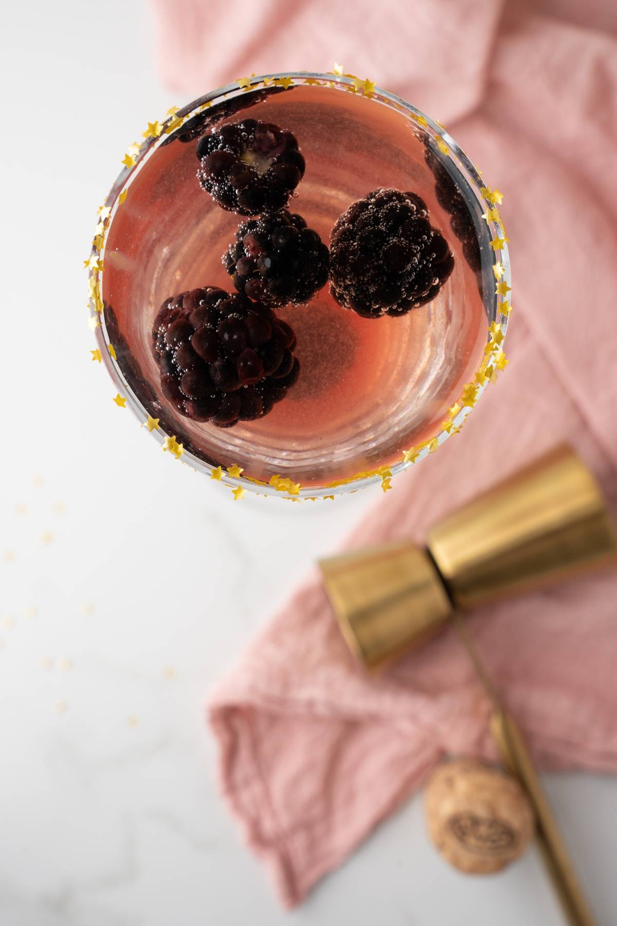 A Lemon Berry Prosecco Cocktail with blackberries and star sprinkles on the rim, served beside a gold jigger and pink cloth.