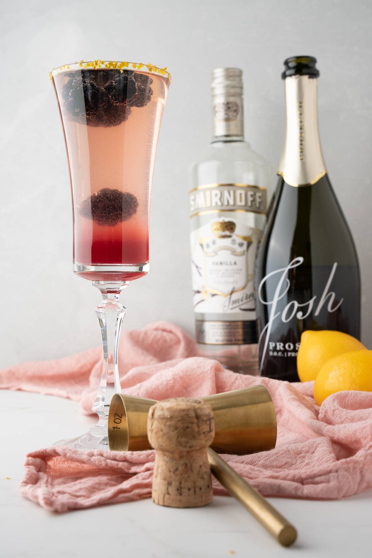 A bubbly Lemon vodka Prosecco Cocktail with blackberries in a flute, featuring Meyer lemon blackberry syrup, surrounded by lemons, prosecco, and bar tools in the background—a simple prosecco drink perfect for any occasion.