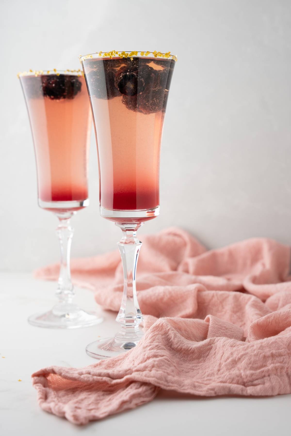 Two tall glasses of pink sparkling Lemon vodka Prosecco Cocktail with berries on top sit beside a soft pink cloth on a white surface.