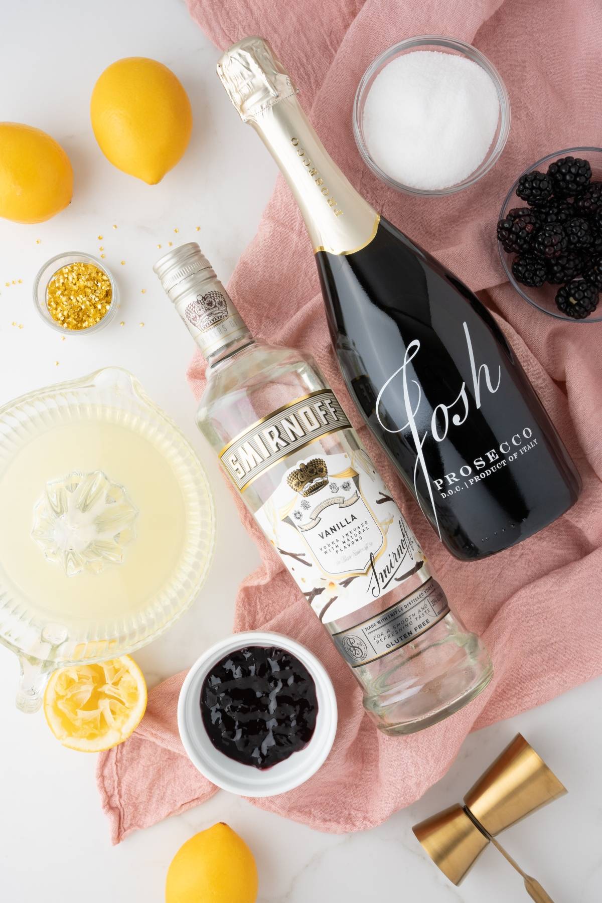 Lemons, sugar, gold sprinkles, blackberries, vodka, and Prosecco are arranged on a pink cloth—perfect for mixing a festive Christmas or New Year’s Eve cocktail with Meyer lemon blackberry syrup.