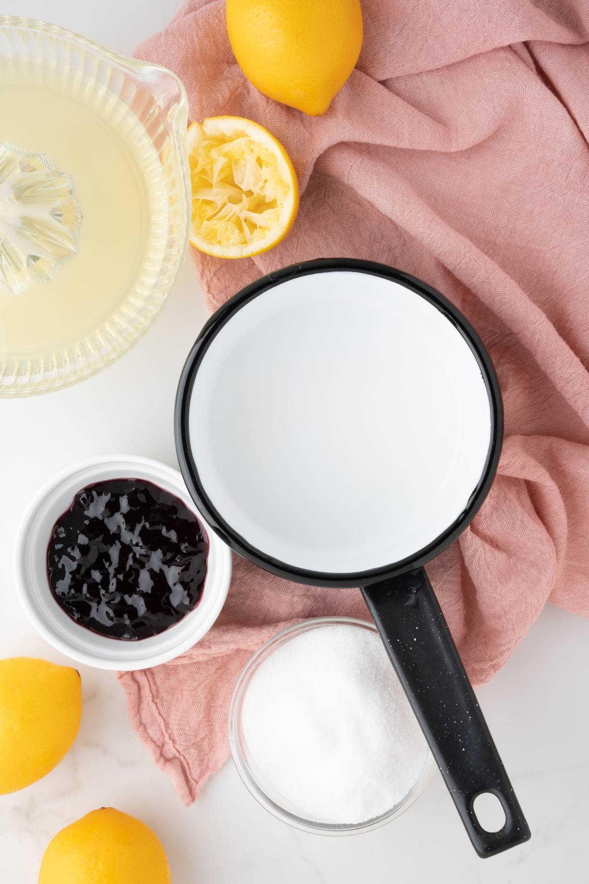 A saucepan, lemons, lemon juice, sugar, and berry jam on a pink cloth and white surface—perfect ingredients for a Lemon Berry Prosecco Cocktail, an easy yet festive simple prosecco drink for Christmas or New Year's Eve.