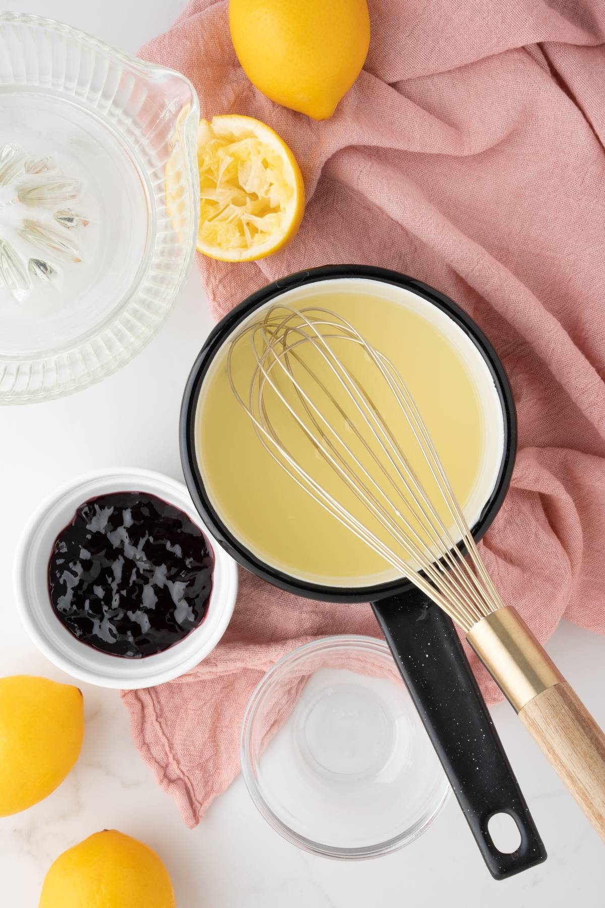 A saucepan with a whisk, lemons, lemon juicer, jam, and a pink cloth on a white surface—perfect for crafting Meyer lemon blackberry syrup for your Lemon Berry Prosecco Cocktail.
