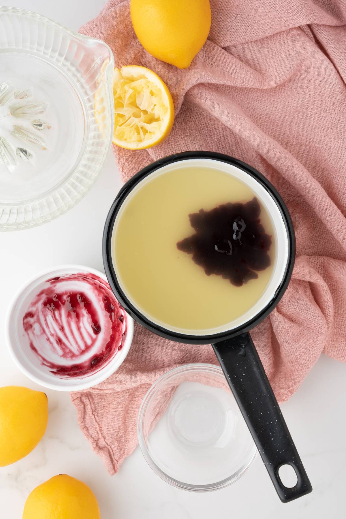 Saucepan with yellow liquid and a dark swirl—perhaps Meyer lemon blackberry syrup—surrounded by lemons, juicer, bowl, and pink cloth on a white surface. Perfect for crafting a refreshing Lemon Berry Prosecco Cocktail.
