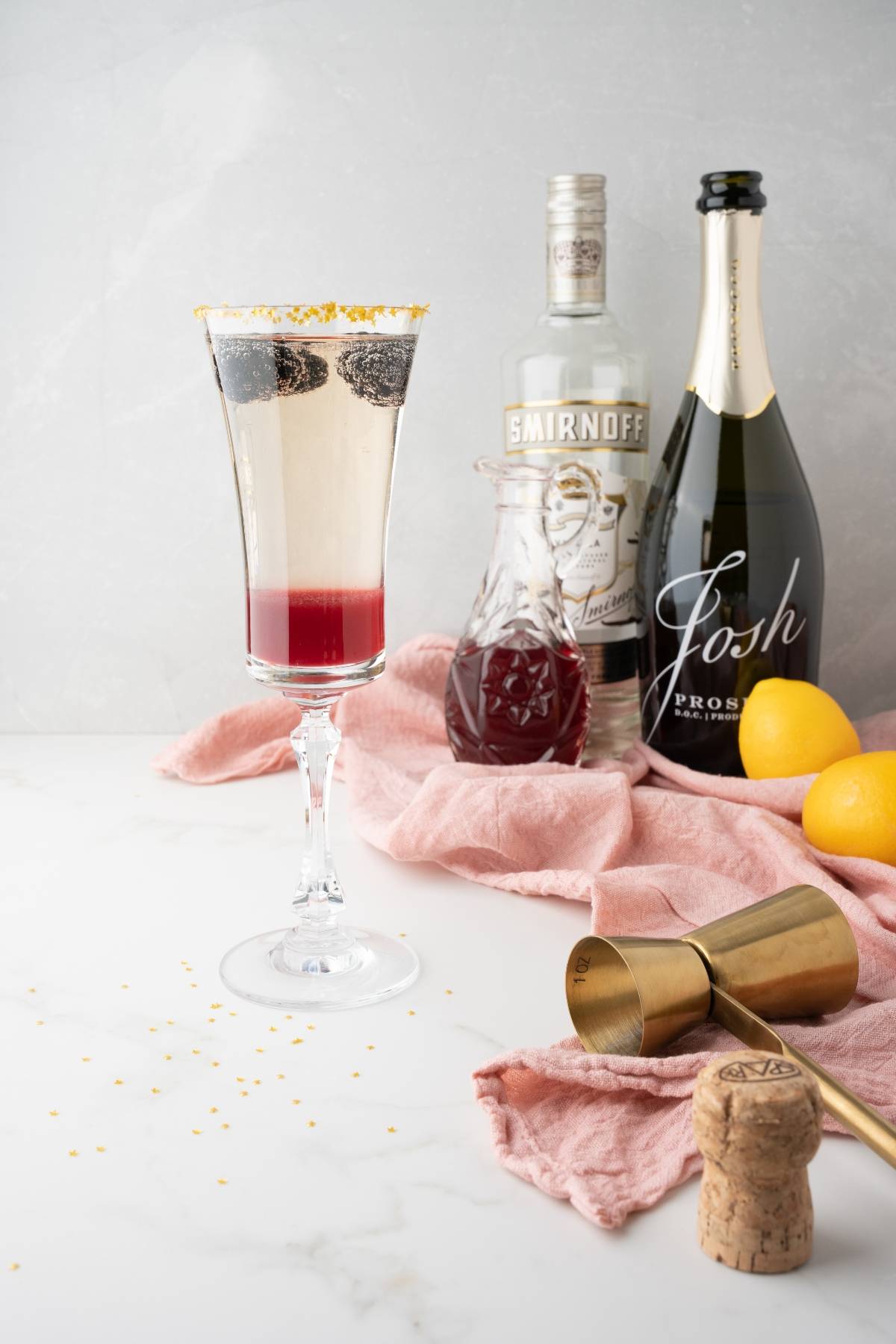 A festive Lemon Vodka Prosecco Cocktail in a flute glass with berries, surrounded by syrup, lemons, and bar tools—a perfect simple prosecco drink for Christmas or New Year’s Eve celebrations.
