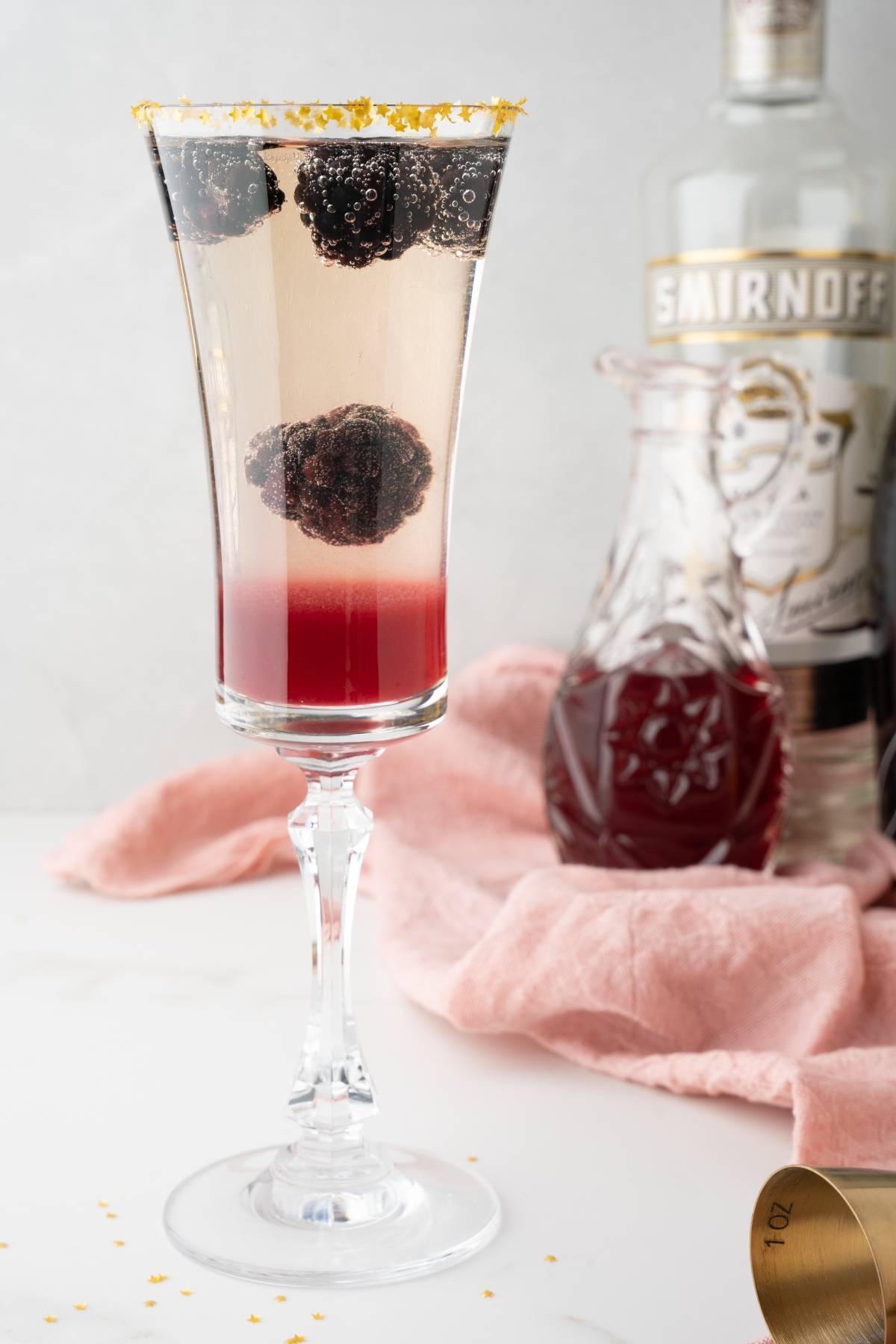 A champagne flute with a layered red and clear Lemon Berry Prosecco Cocktail, blackberries, and a sugared rim, with a vodka bottle behind—perfect as a festive Christmas or New Year’s Eve cocktail.