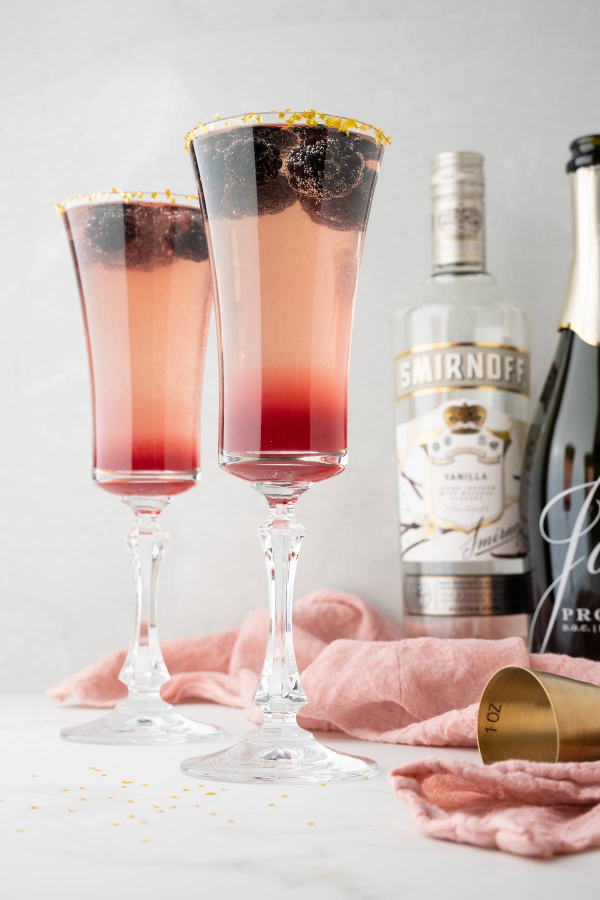 Two tall glasses of Lemon Berry Prosecco Cocktail, beautifully layered with pink hues and blackberries, sit beside bottles of Smirnoff vodka and Prosecco—a simple prosecco drink perfect for any celebration.