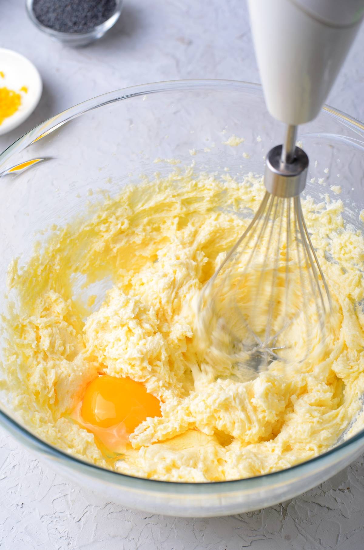 Hand mixer whisking egg into creamed butter and sugar for a lemon poppy seed loaf, with ingredients nearby in a glass bowl.