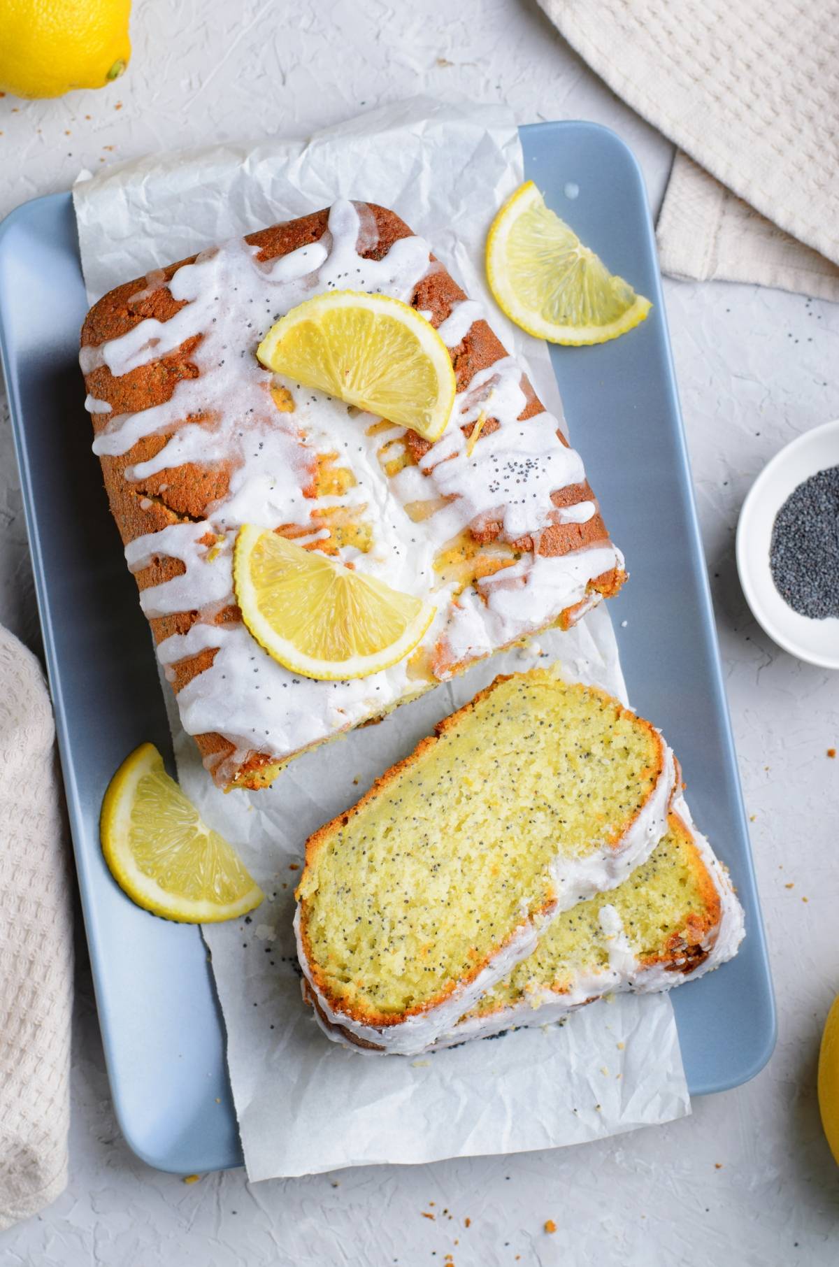 Lemon poppy seed loaf with a sweet icing, garnished with lemon slices, sits on a blue tray beside a bowl of poppy seeds—perfect for citrus lovers.
