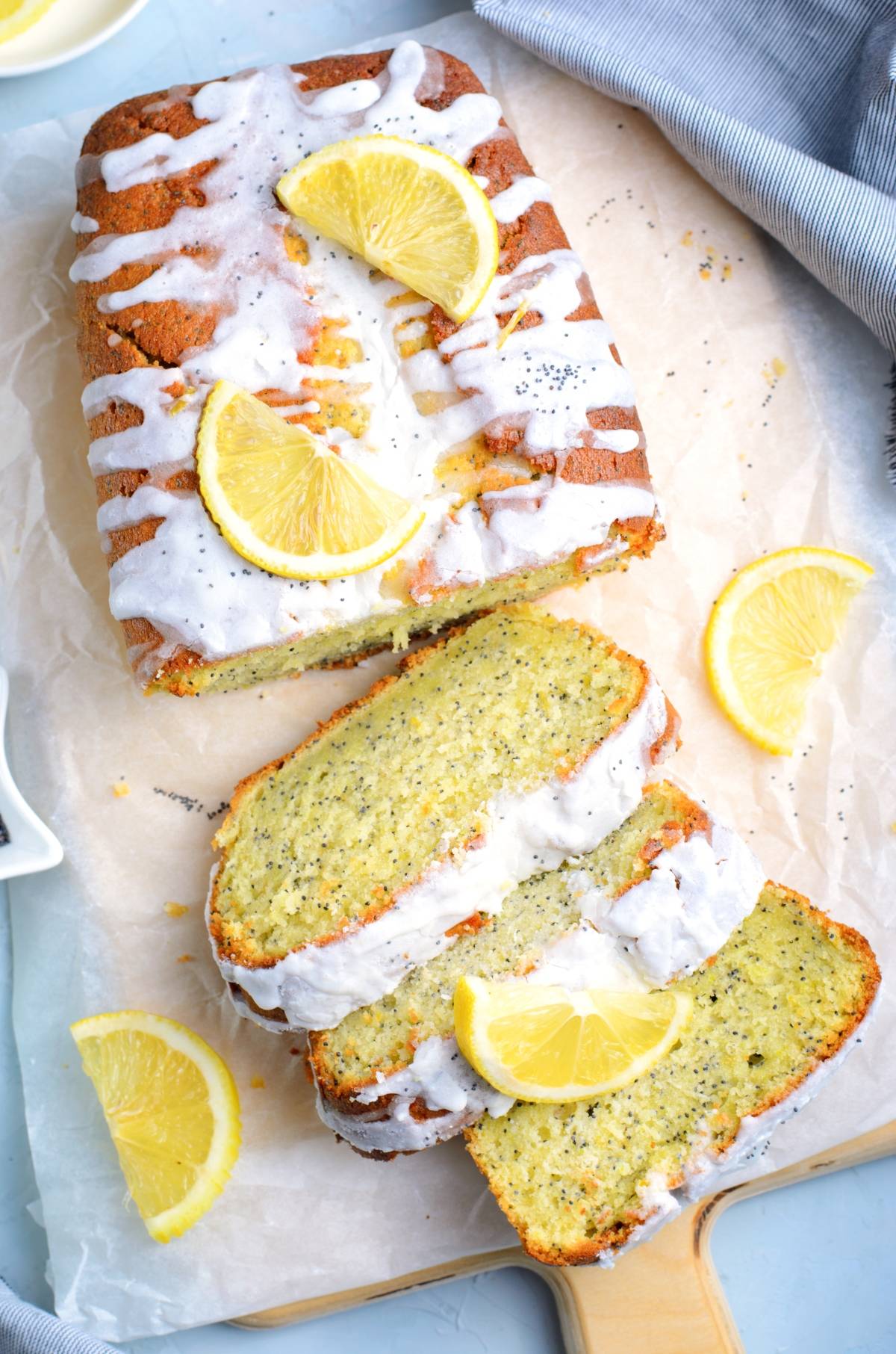 A freshly baked lemon poppy seed loaf with white icing and bright lemon slices, partially sliced on parchment paper.
