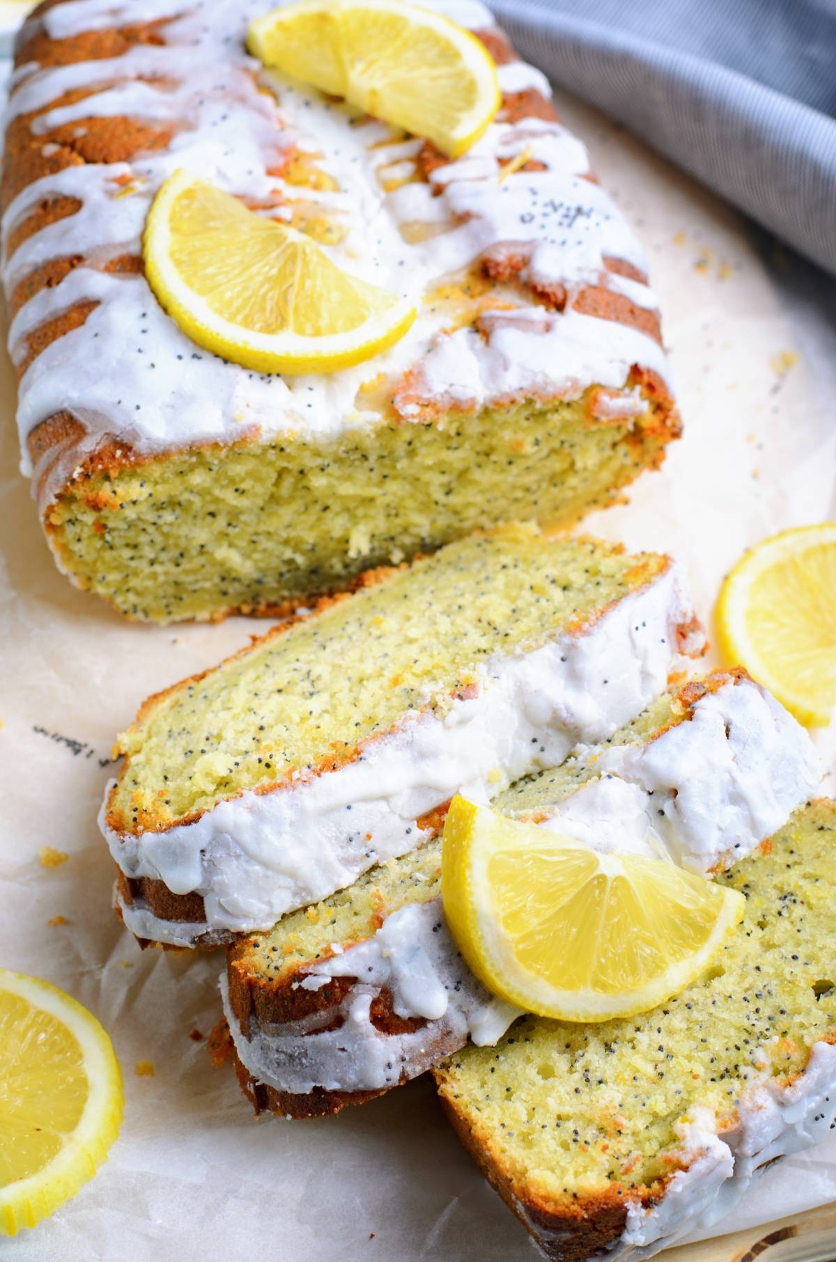 Sliced lemon poppy seed loaf with white icing and lemon wedges on top, this beautiful lemon poppy seed loaf rests on parchment paper.