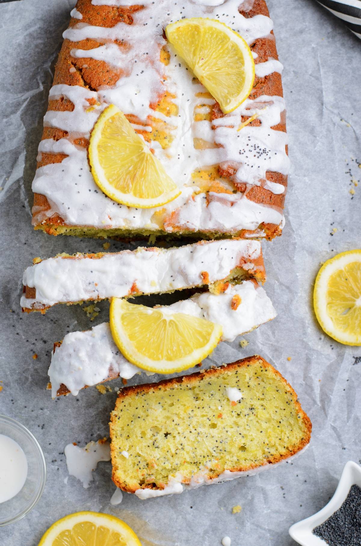 Moist lemon poppy seed loaf with sweet icing and fresh lemon slices, partially sliced on parchment paper.