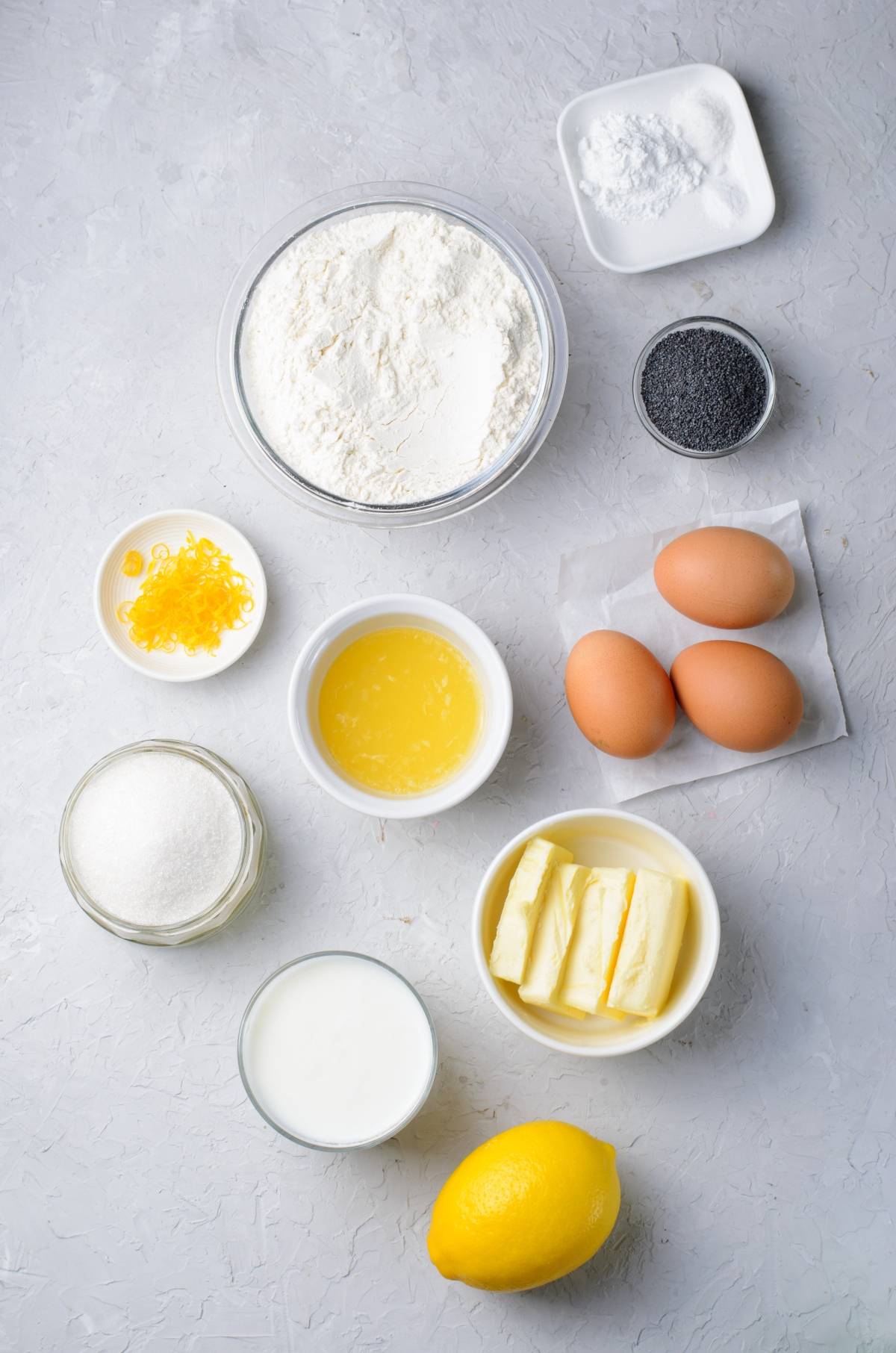 Baking ingredients arranged on a light surface for a lemon poppy seed loaf: flour, eggs, butter, lemon, sugar, milk, poppy seeds, zest, and salt.