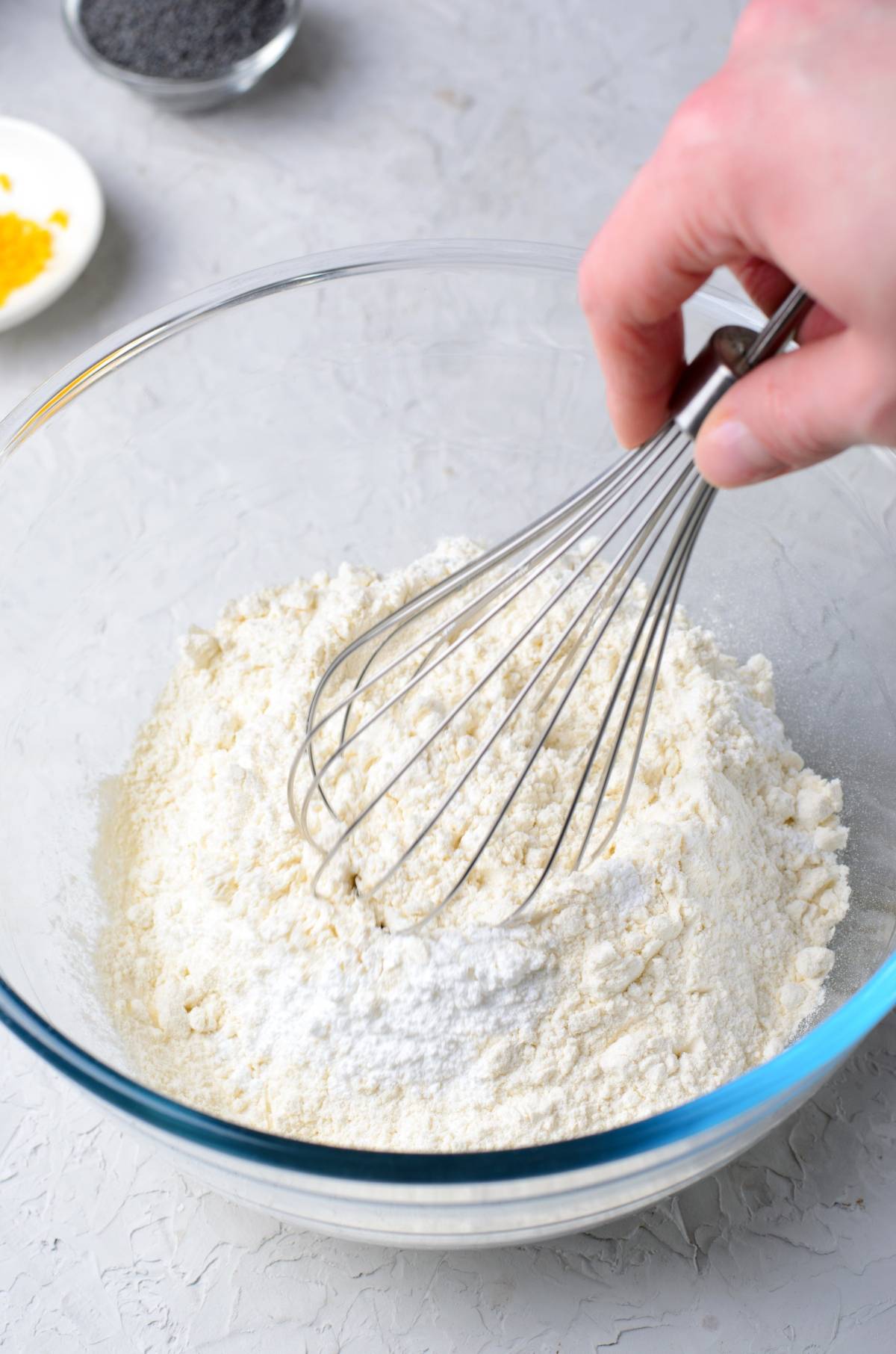 Hand whisking flour in a glass bowl on a light surface, with ingredients for a lemon poppy seed loaf visible in the background.
