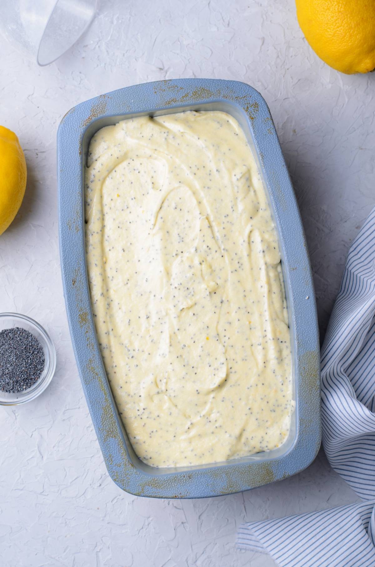 Rectangular baking pan filled with lemon poppy seed loaf batter, surrounded by fresh lemons and a bowl of poppy seeds.