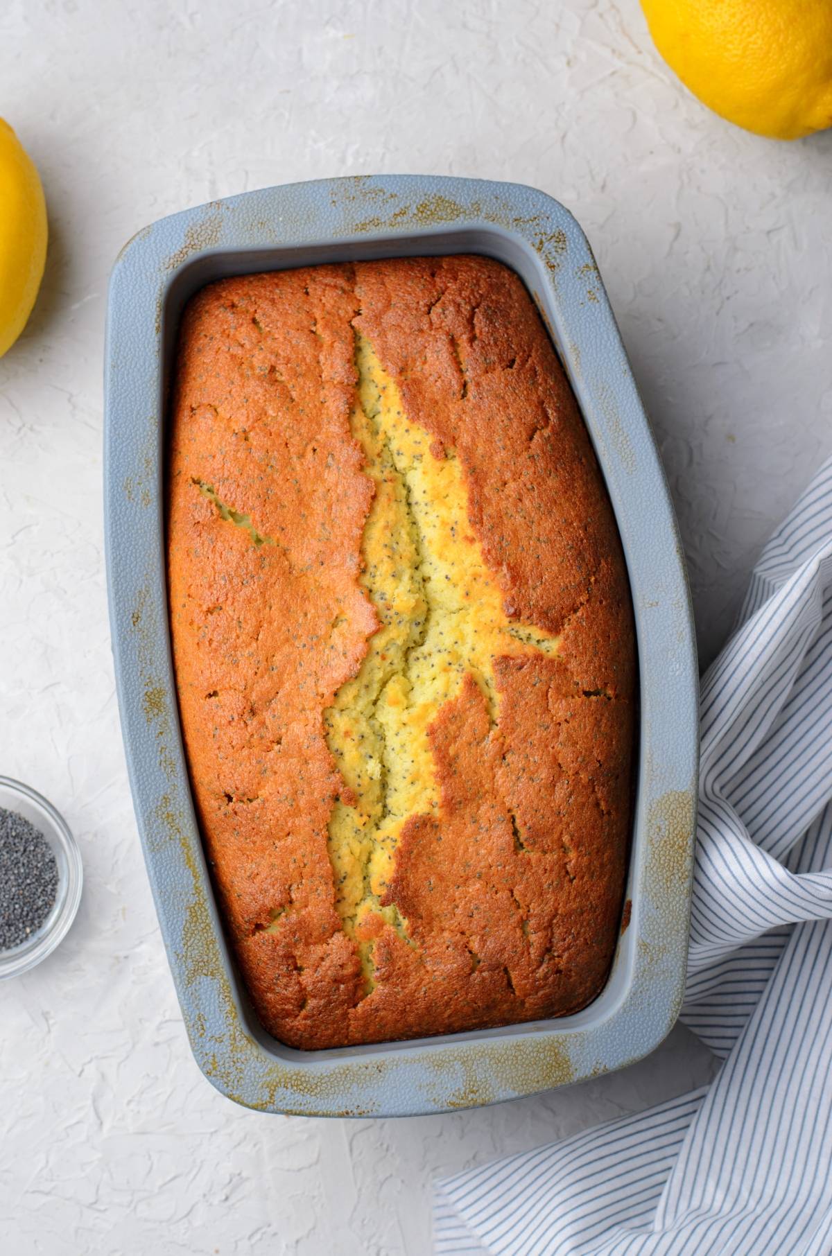 Golden lemon poppy seed loaf in a gray baking pan, its cracked top revealing a tender crumb, sits beside fresh lemons and a striped cloth.