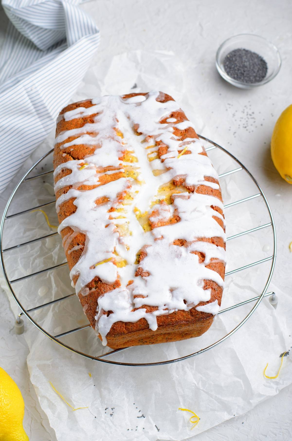 Iced lemon poppy seed loaf sits on a cooling rack with lemons and poppy seeds nearby on a white surface.