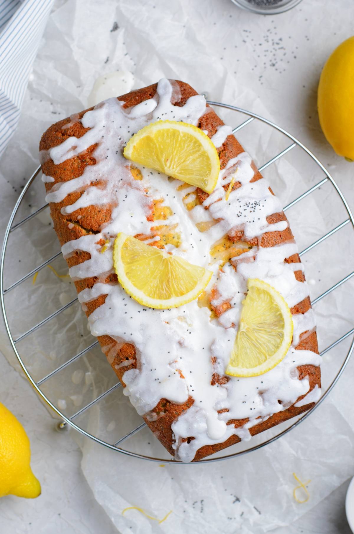 Lemon poppy seed loaf cake with white icing, lemon slices, and poppy seeds on a cooling rack, surrounded by fresh lemons.