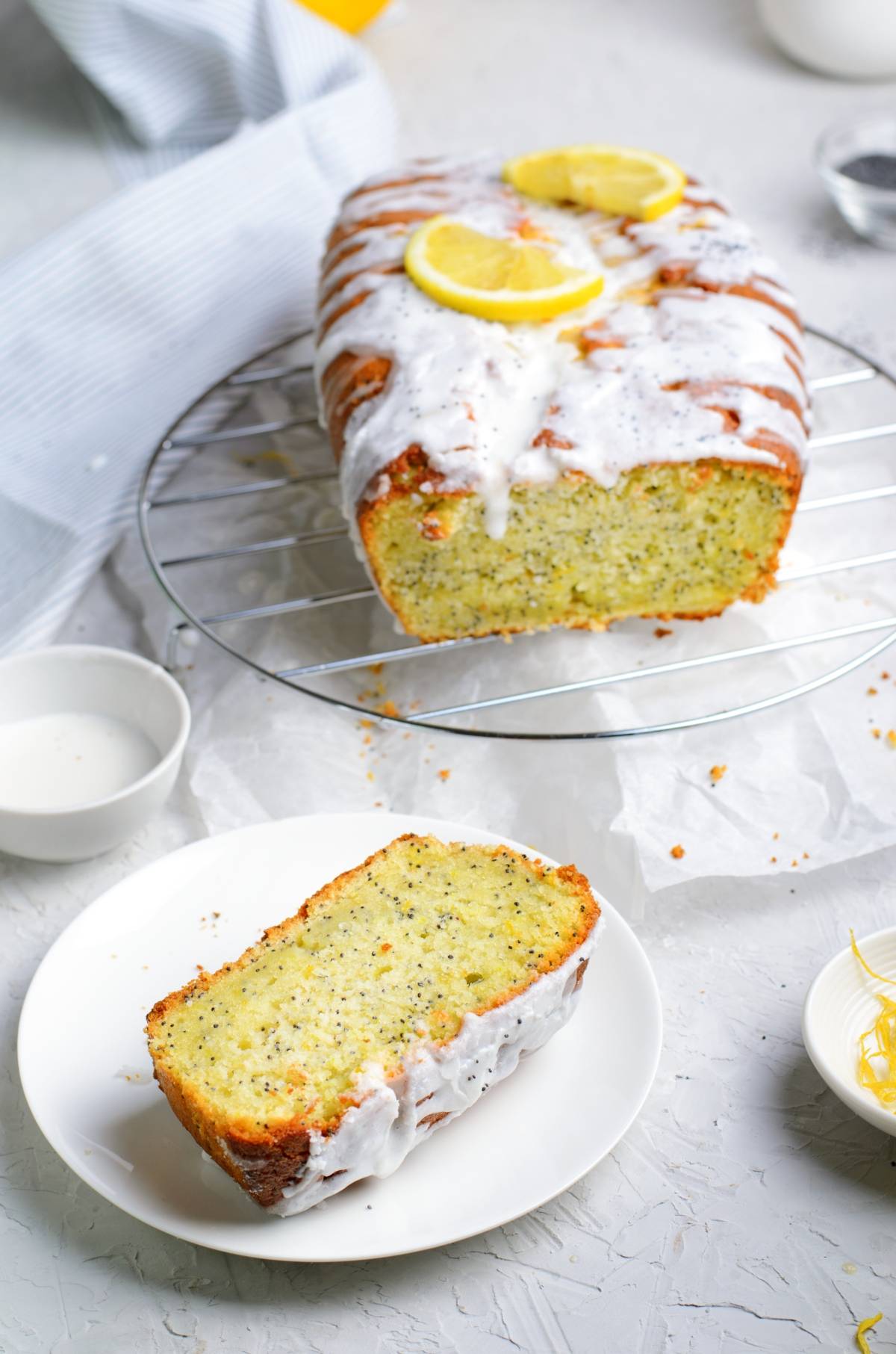 A slice of lemon poppy seed loaf with icing rests on a plate, while the freshly baked lemon poppy seed loaf sits on a cooling rack, topped with vibrant lemon slices.