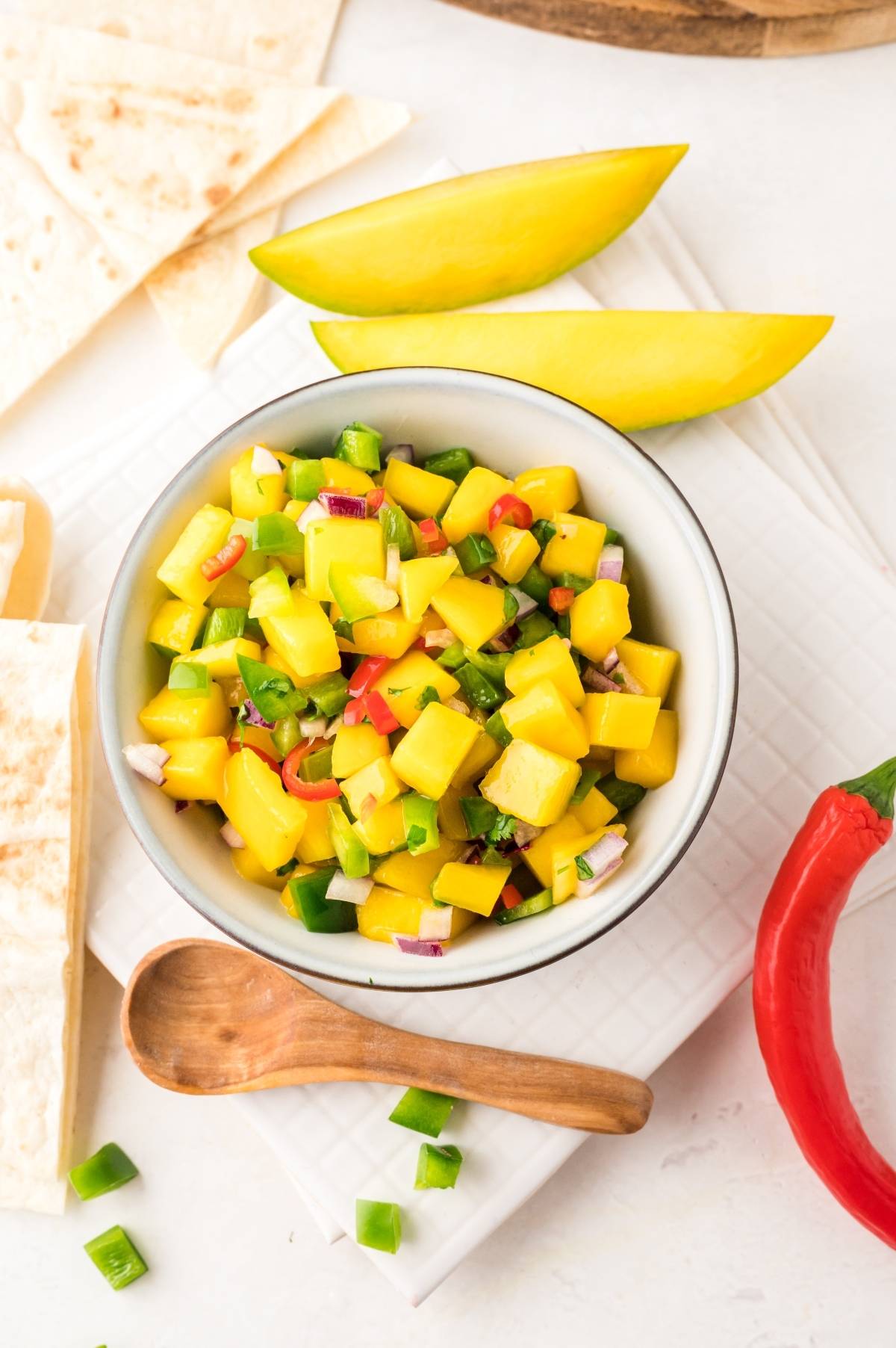 Bowl of salsa made with fresh mangoes, diced vegetables, wooden spoon, tortilla slices, and mango wedges nearby.