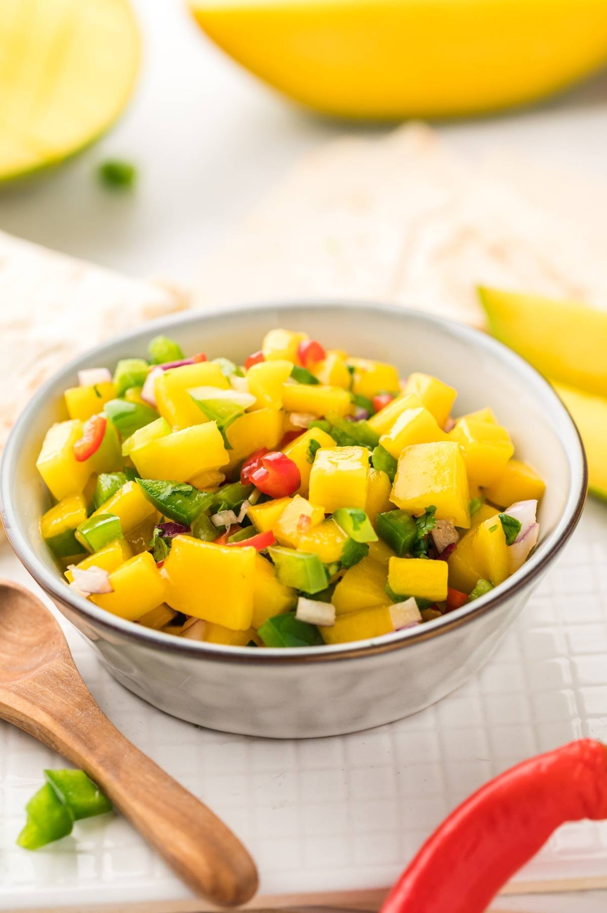 A bowl of Mexican fruit salsa made with fresh mangoes, chopped peppers, onions, and herbs sits beside a wooden spoon.