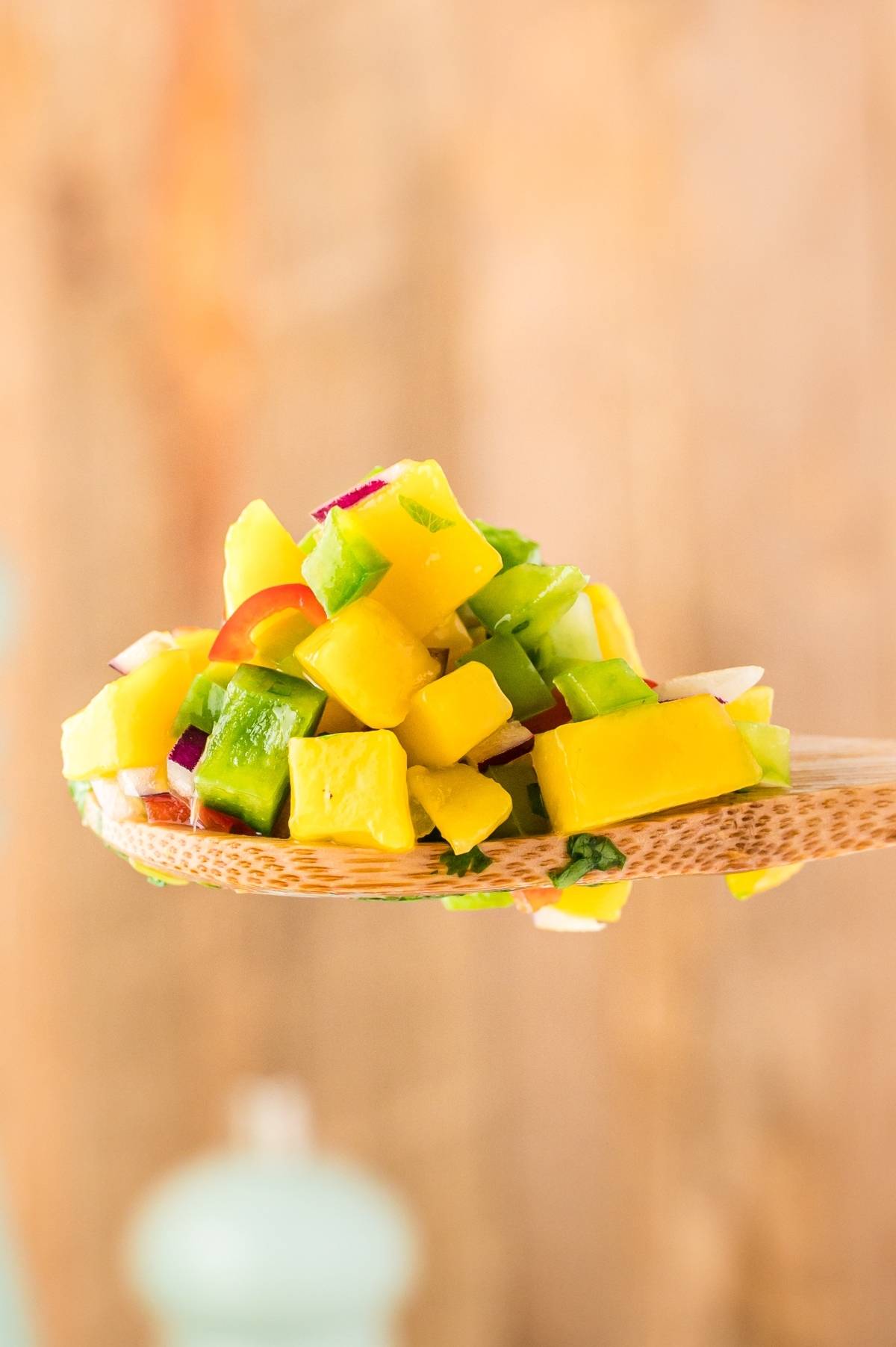 A wooden spoon holds a vibrant mango salsa, featuring diced mango, cucumber, and red onion—a perfect summer salsa recipe.