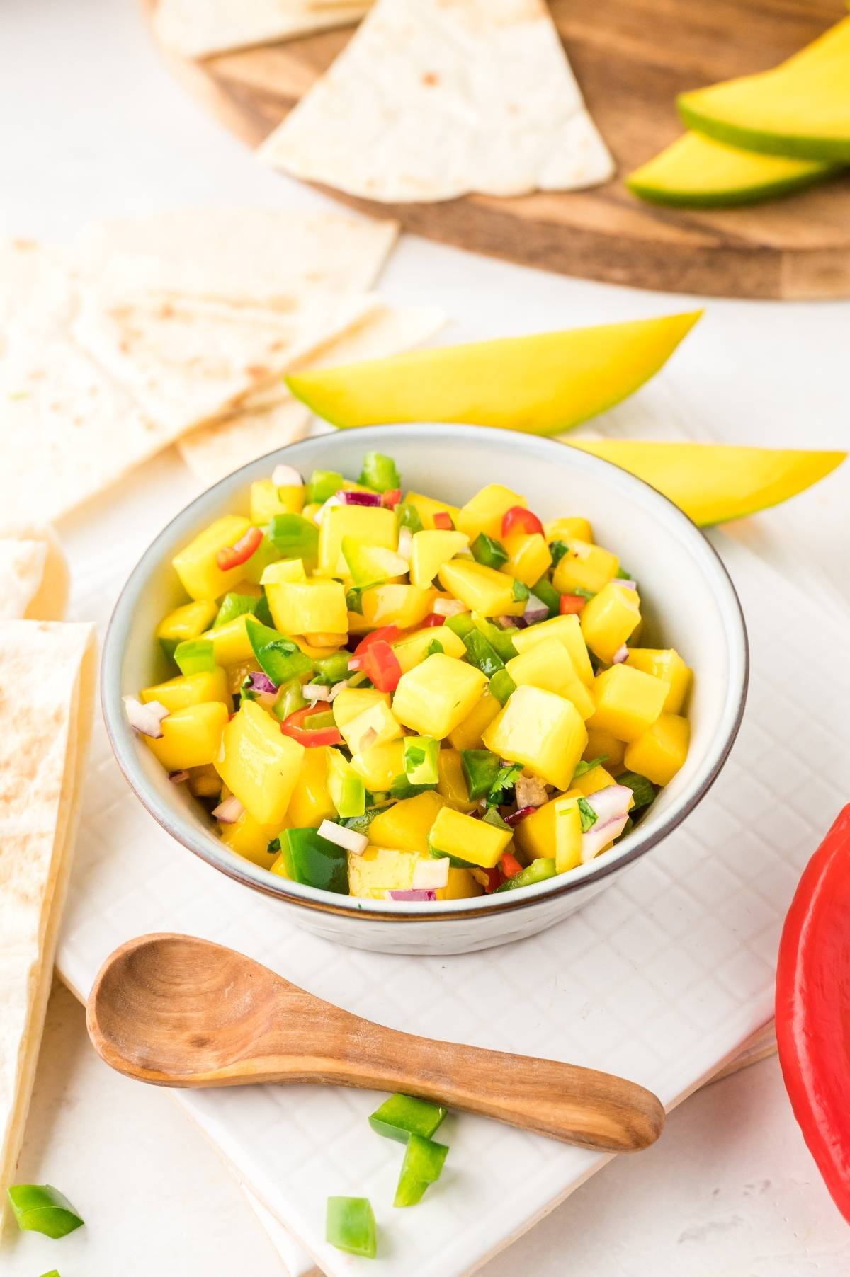 A bowl of mango salsa, a summer salsa recipe with chopped peppers and onions, is surrounded by tortilla chips and fresh mango slices.
