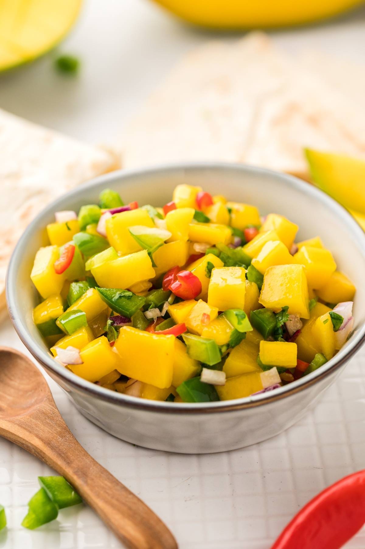 A bowl of fresh Mexican fruit salsa with diced mango, red onion, peppers, and herbs sits next to a wooden spoon.