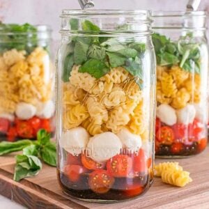 Three mason jar Caprese pasta salads layered with mozzarella, cherry tomatoes, greens, and dressing are placed on a wooden board.