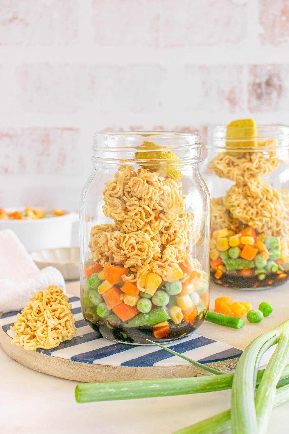 Two jars filled with dry ramen noodles and mixed frozen vegetables, placed on a striped serving board.