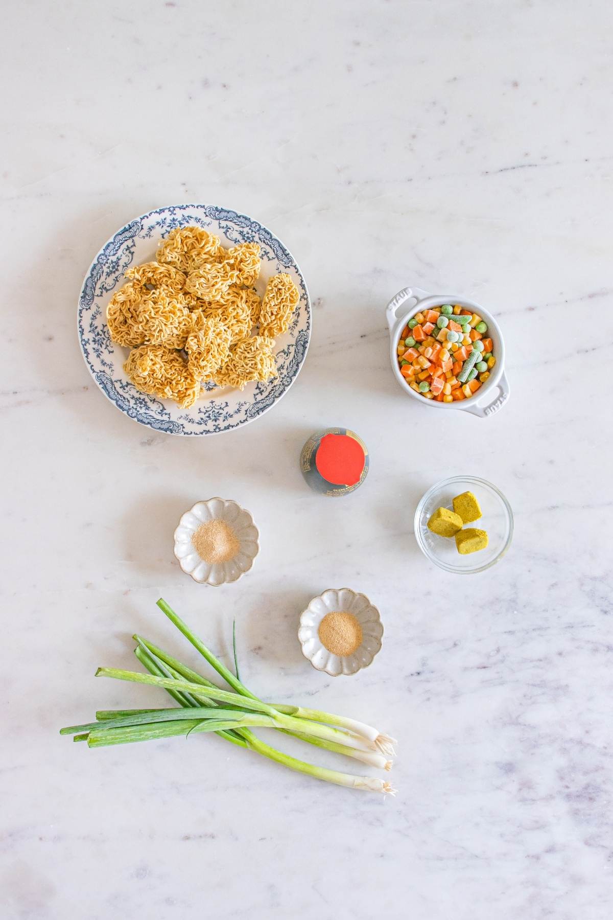 Uncooked ramen noodles, frozen mixed veggies, green onions, bouillon cubes, soy sauce, and spices on a countertop.