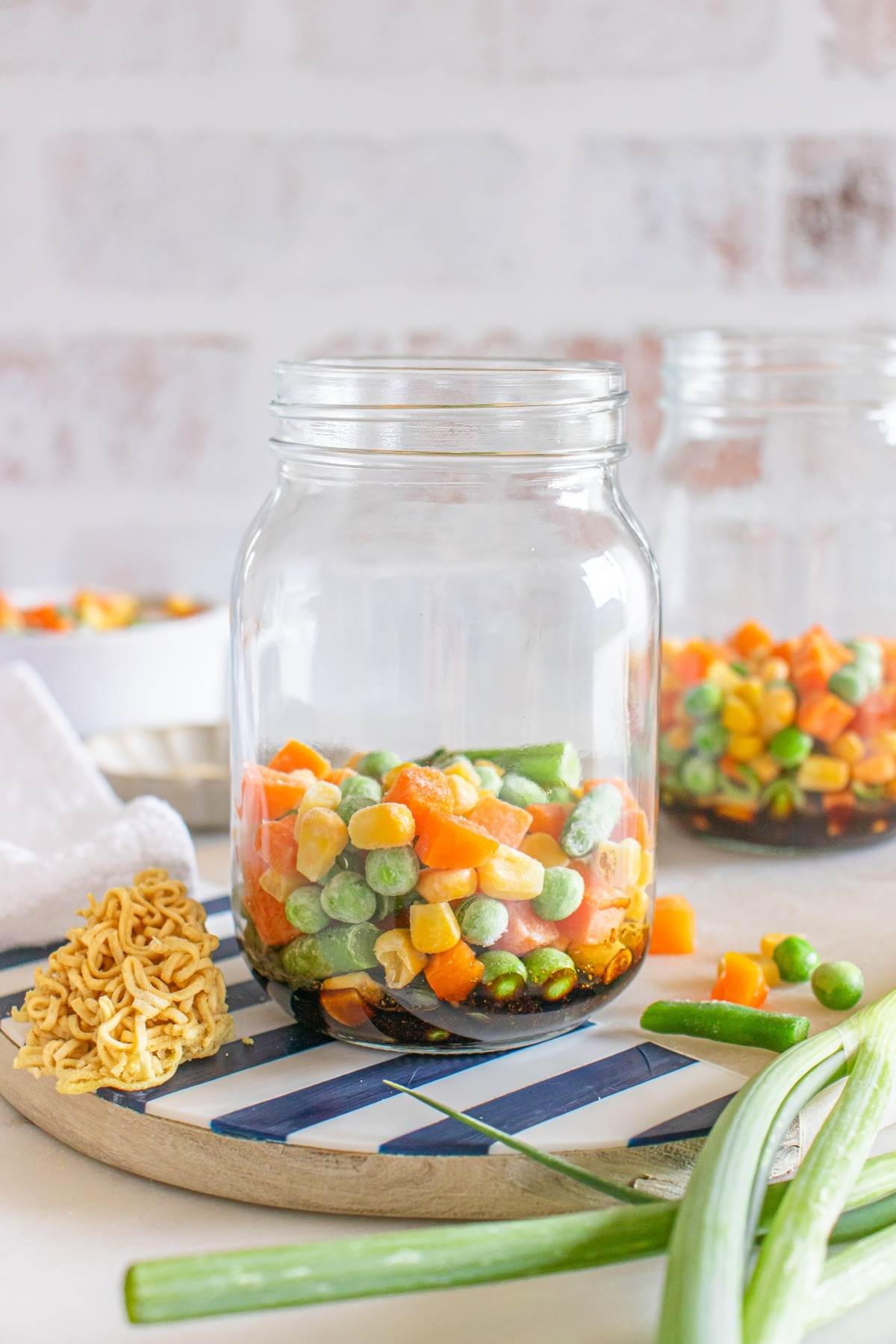 A mason jar filled with frozen mixed vegetables sits on a tray beside uncooked ramen noodles and green onions.