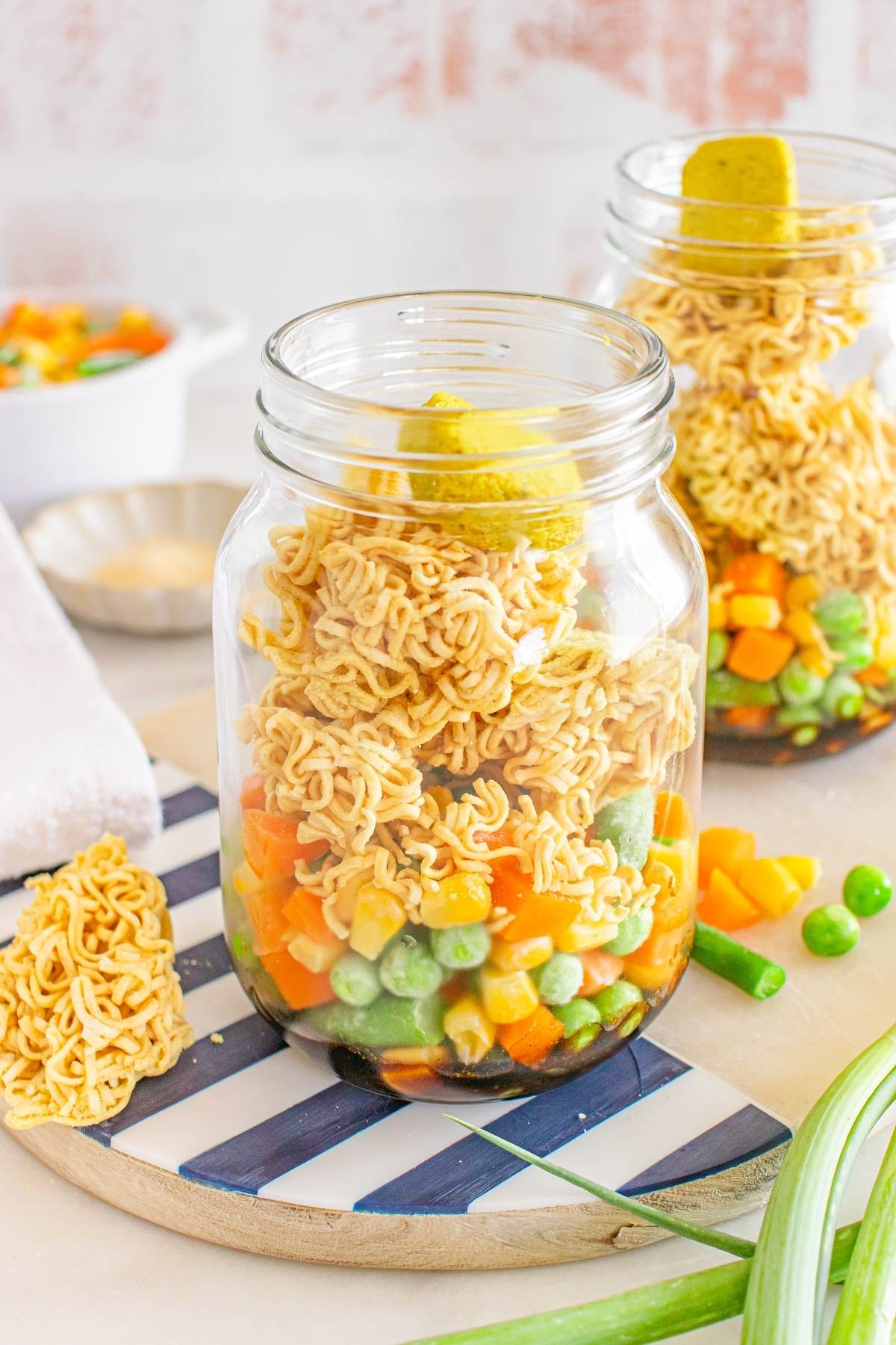 A mason jar filled with dry noodles, frozen mixed vegetables, and a bouillon cube on top.