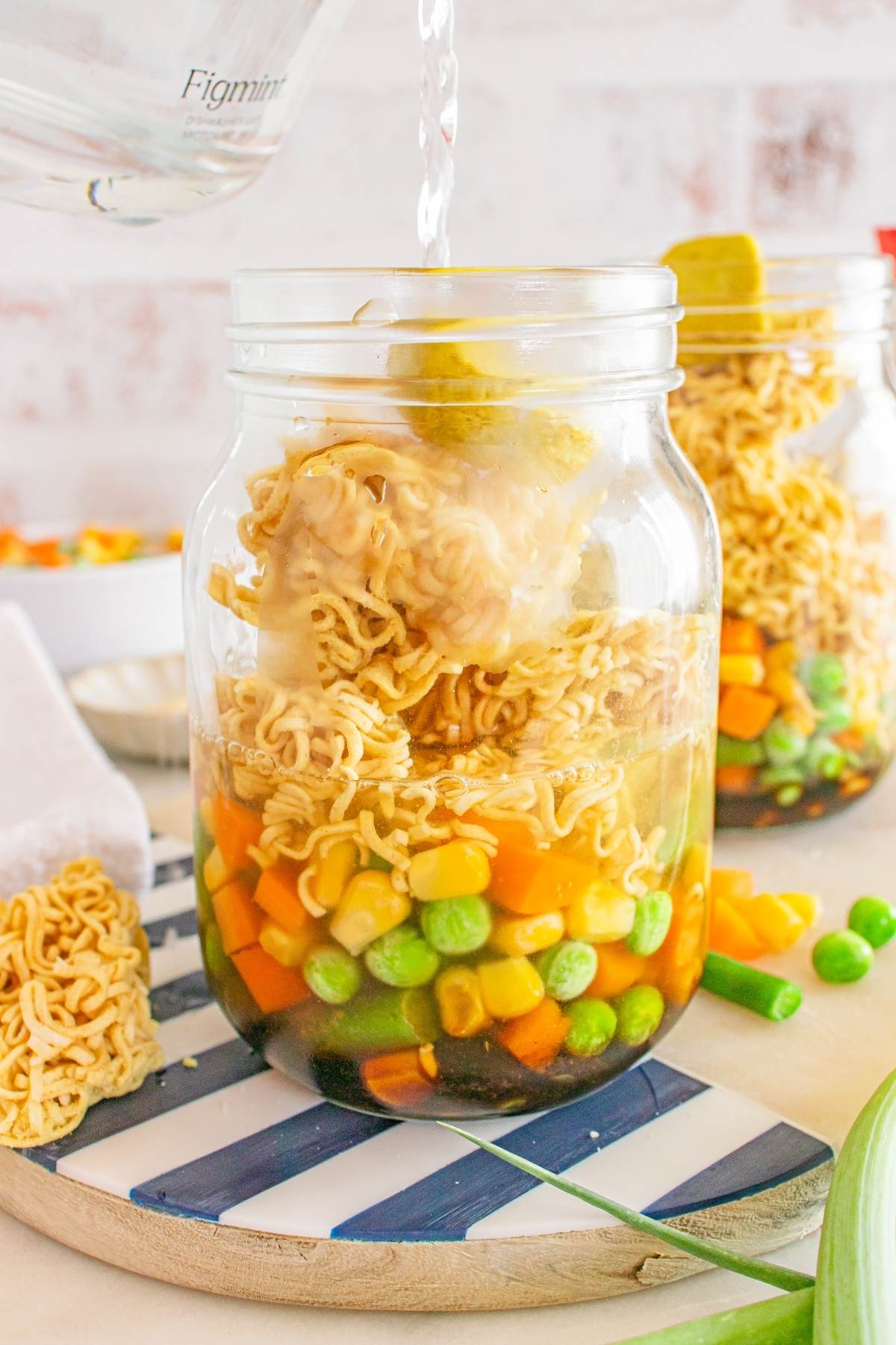 Clear jars filled with dried ramen noodles and mixed vegetables being topped with hot water.