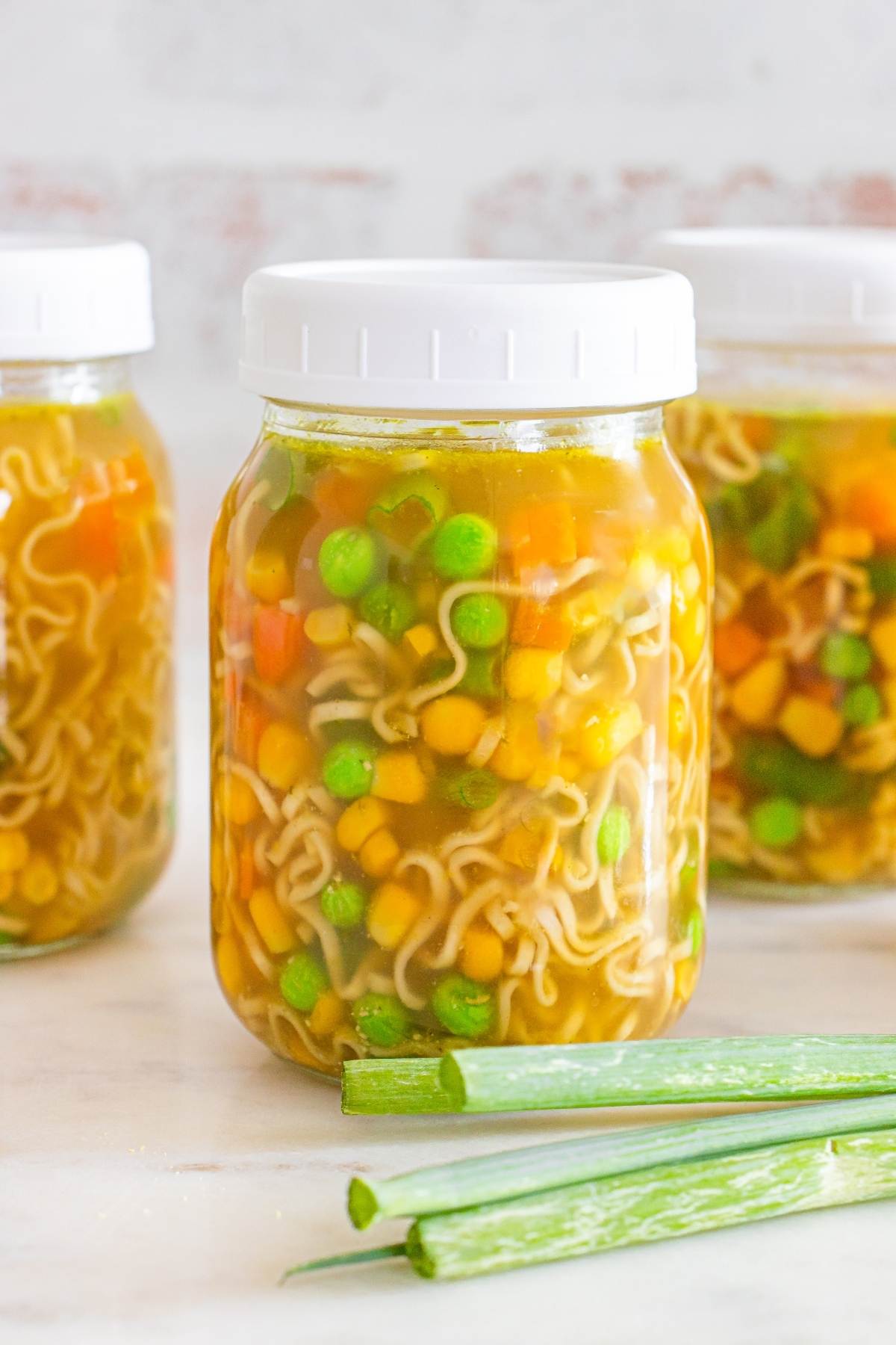 Three jars of vegetable noodle soup with white lids, with green onions in front on a light surface.