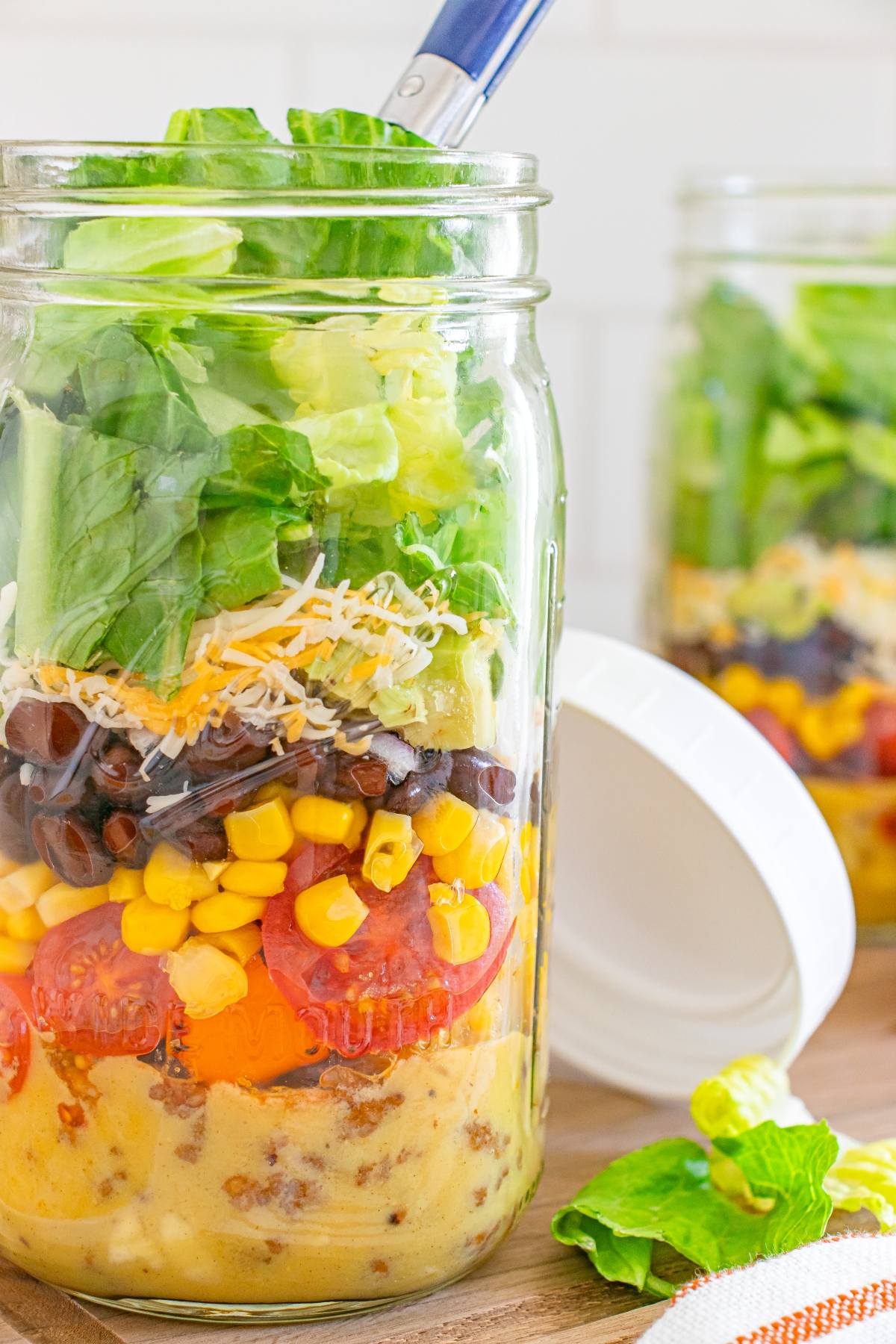 A mason jar filled with layered salad ingredients including dressing, tomatoes, corn, beans, cheese, and lettuce.
