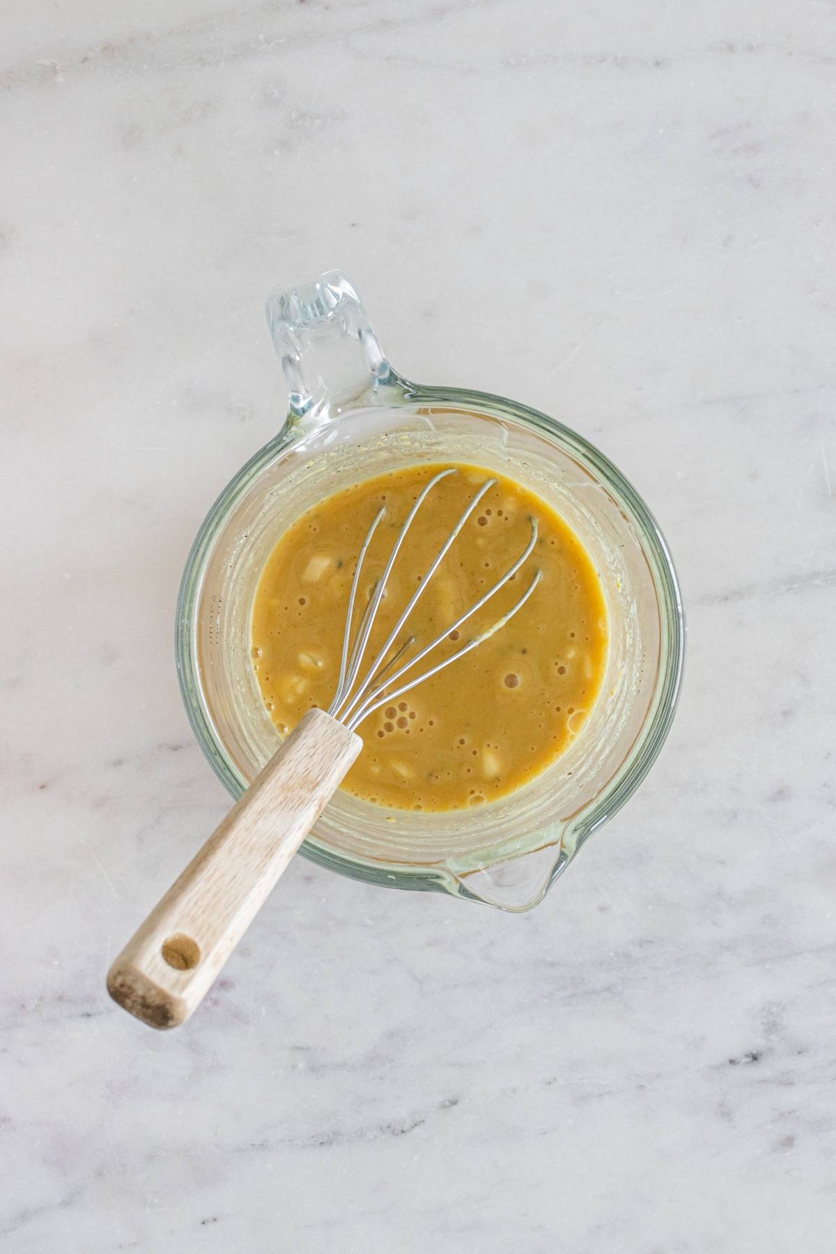 Glass measuring cup with a metal whisk mixing a yellow batter on a light marble countertop.