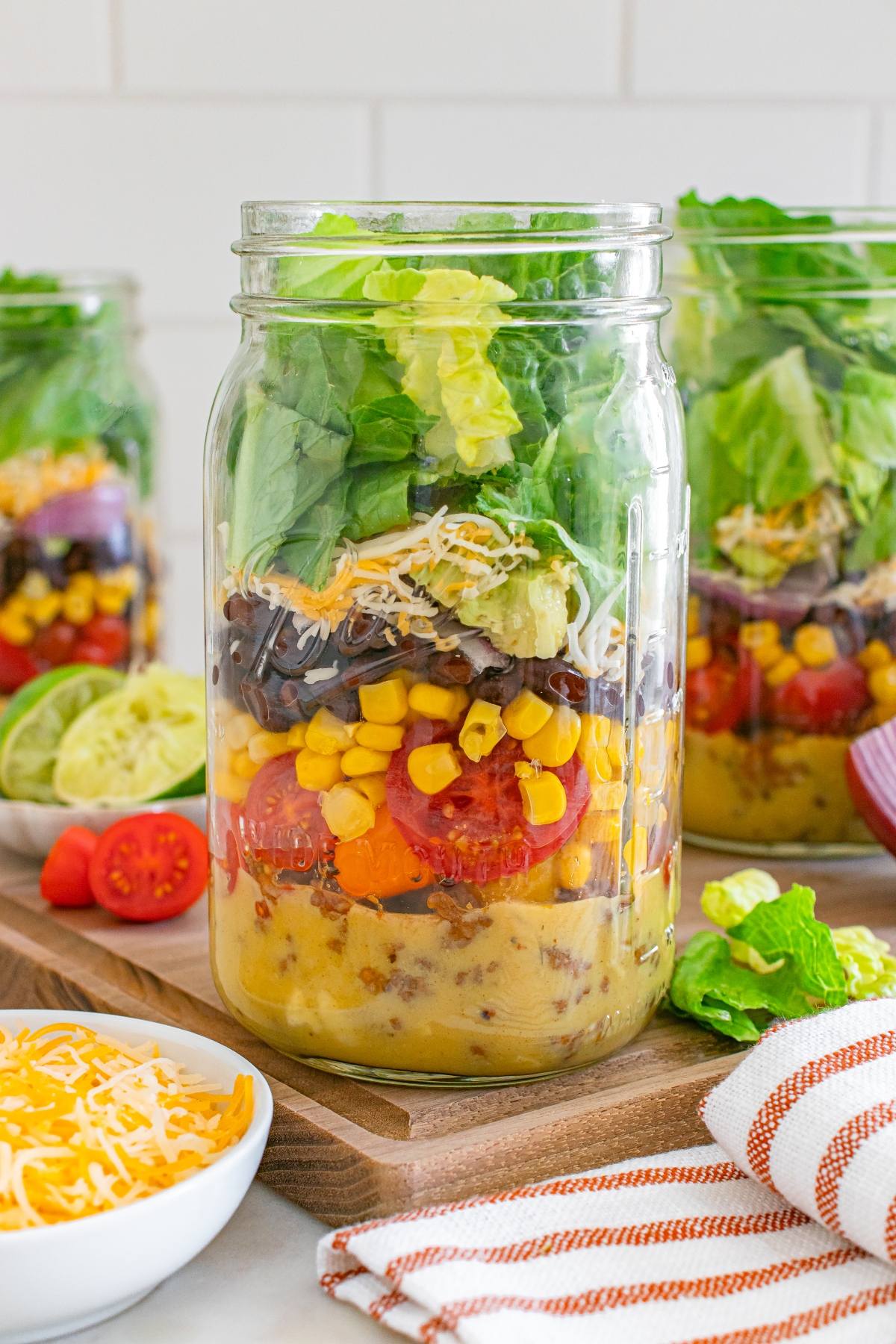Mason jar salad with layers of dressing, corn, tomatoes, black beans, cheese, and lettuce on a wooden board.