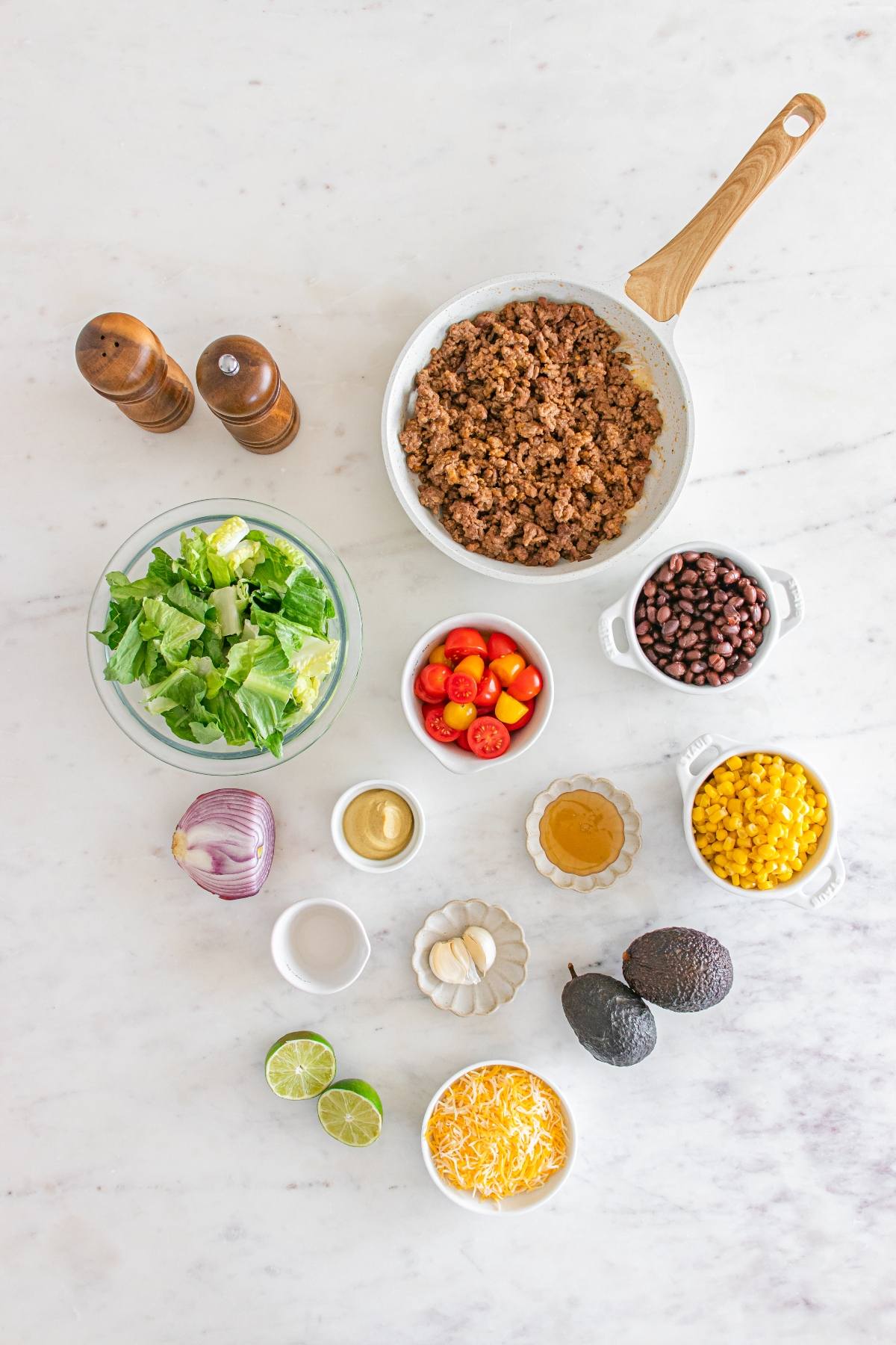Ingredients for taco salad, including ground beef, lettuce, beans, corn, avocado, cheese, lime, and seasonings.