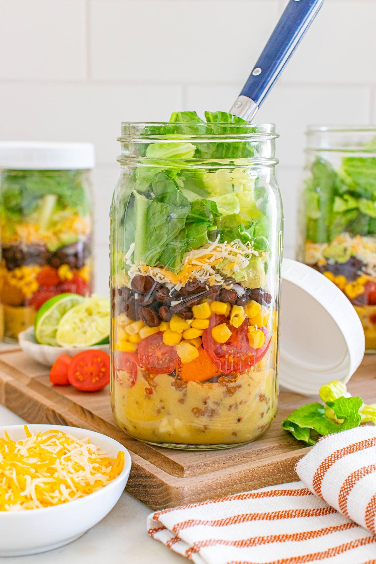 A salad jar layered with dressing, tomatoes, corn, beans, cheese, and lettuce sits on a wooden board.