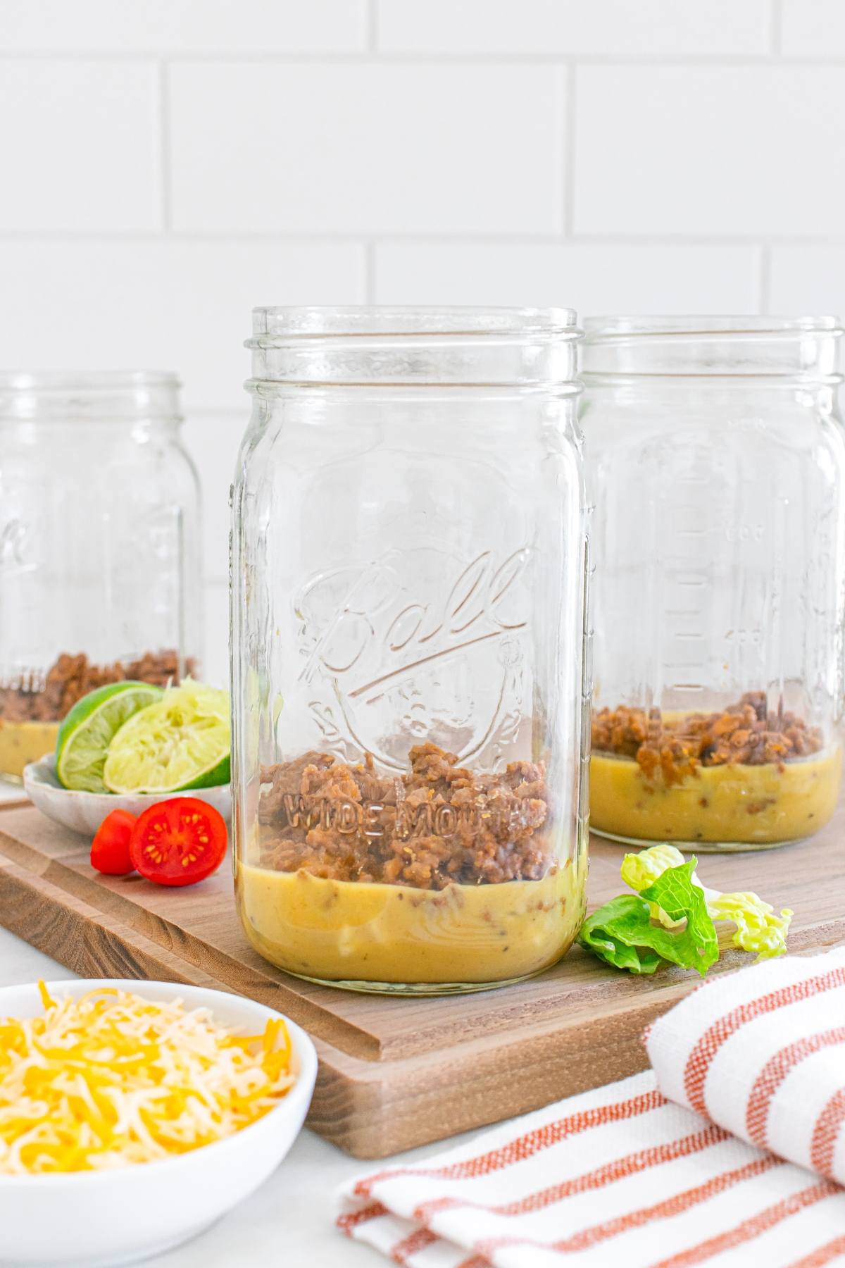 Three glass jars with layers of chili and cheese, surrounded by limes, lettuce, sliced chili, and shredded cheese.