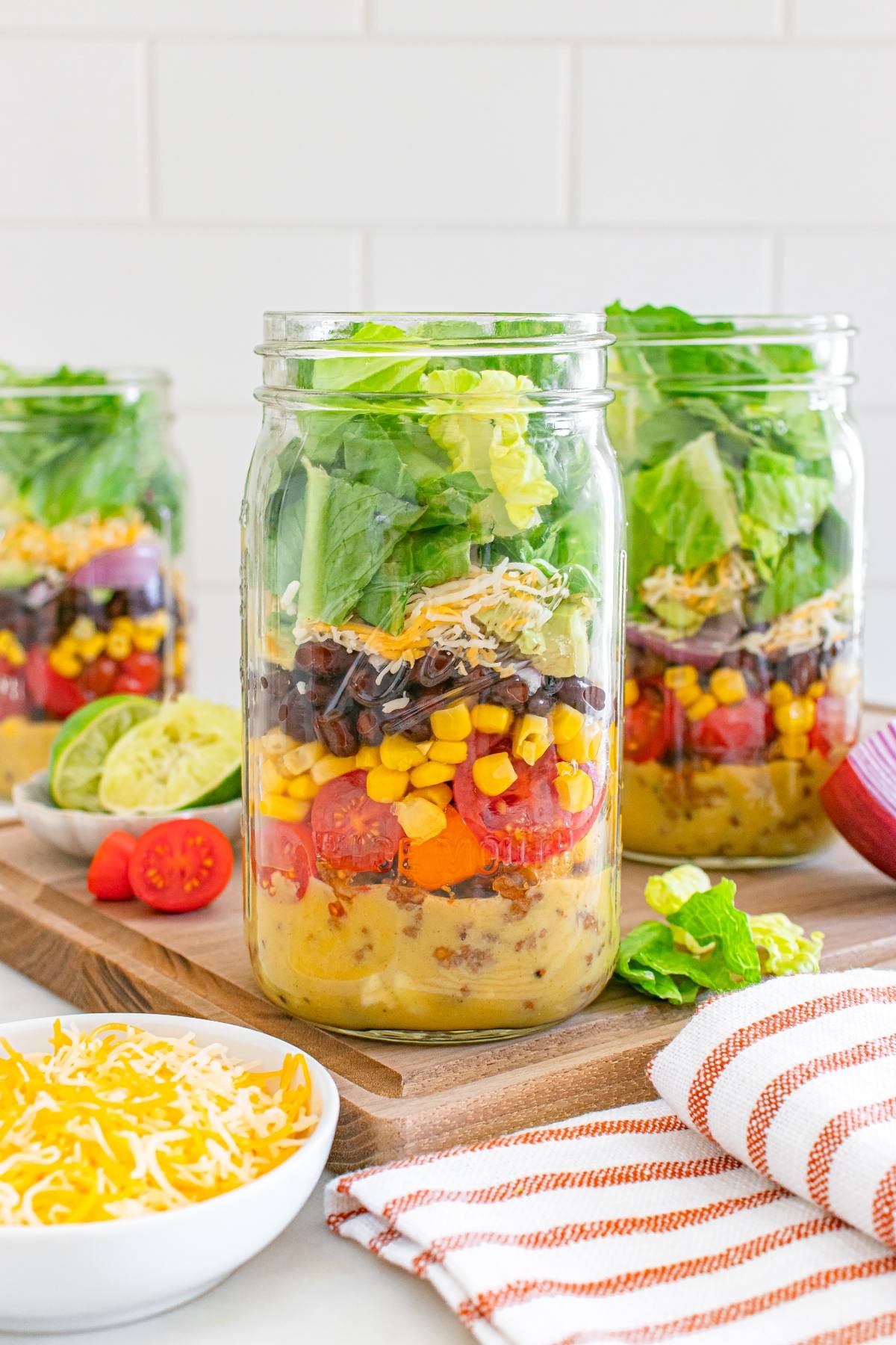Three mason jars layered with salad ingredients, including lettuce, beans, corn, tomatoes, and cheese.