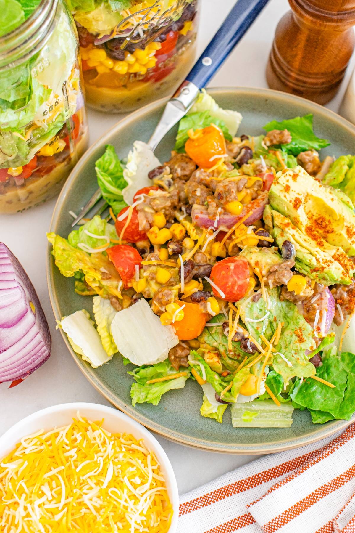 A plate of taco salad with lettuce, tomatoes, beans, corn, avocado, and shredded cheese. Jars of salad nearby.