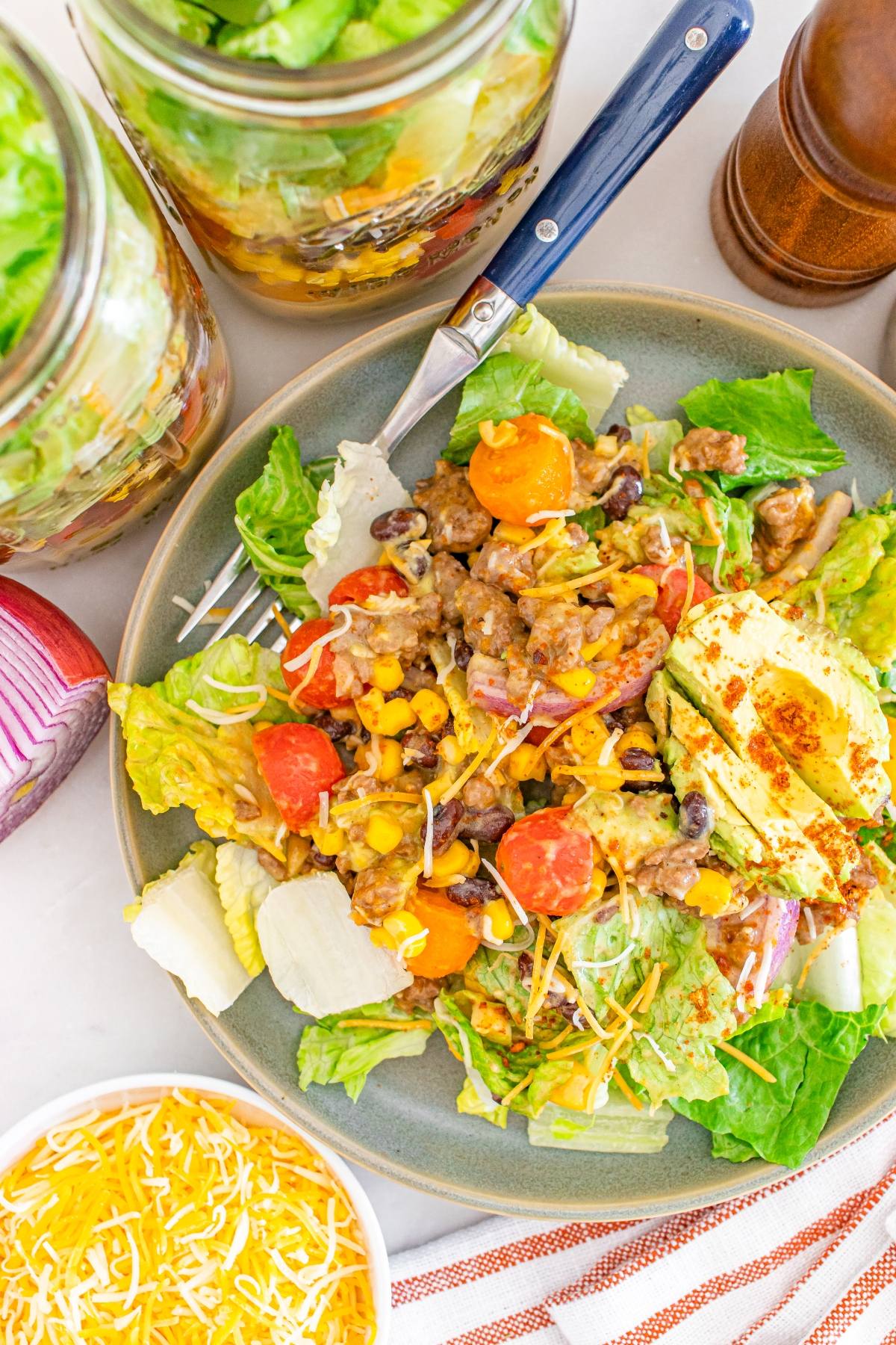 Taco salad with lettuce, cherry tomatoes, corn, ground meat, avocado, and shredded cheese on a plate.