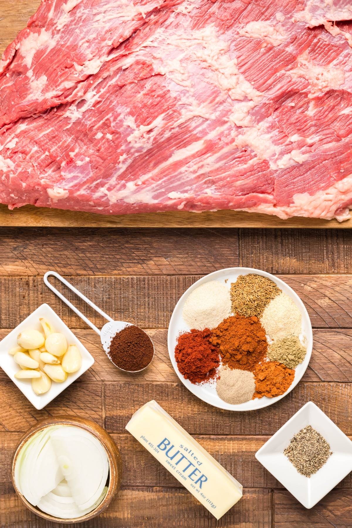 Raw brisket with spices, garlic, onion, butter, and pepper arranged on a wooden surface—perfect ingredients for an easy Texas smoked brisket recipe.