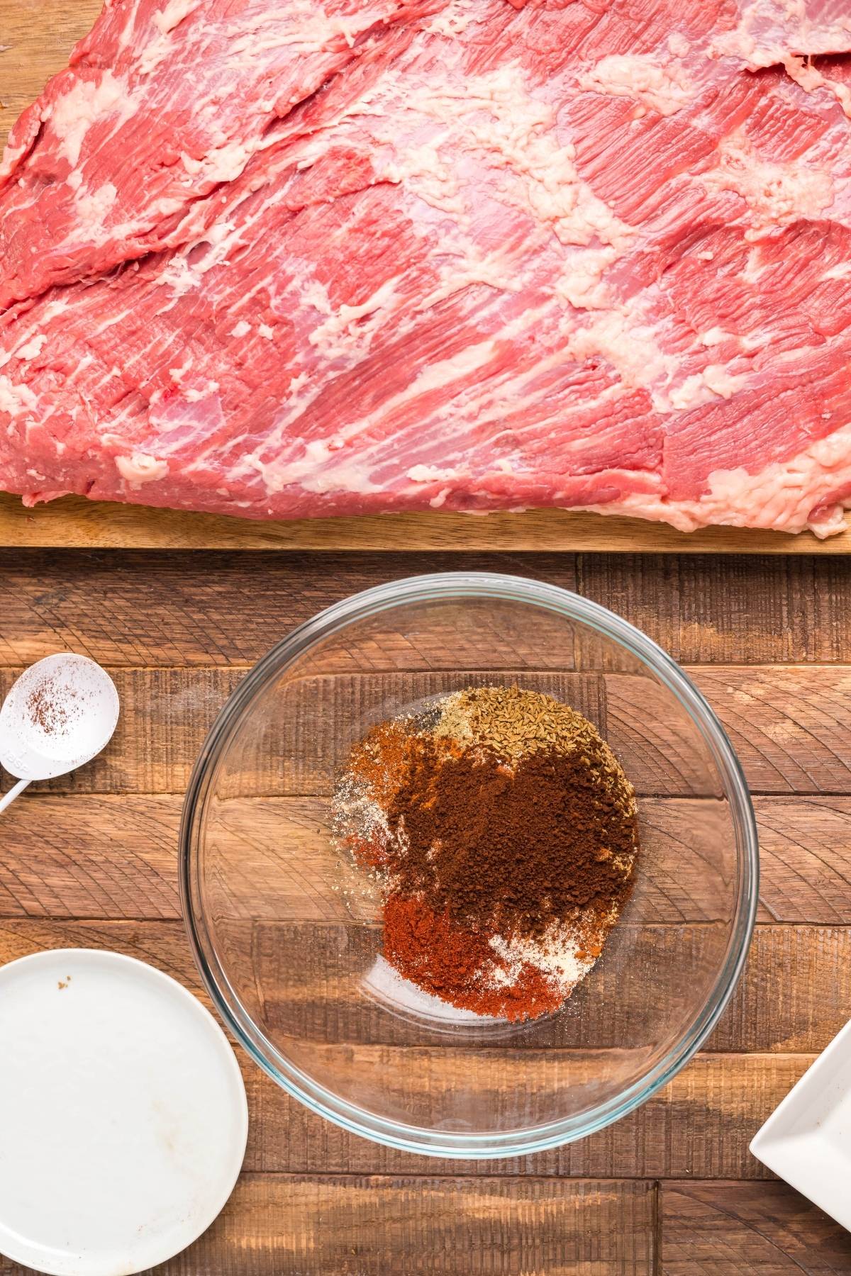 Raw brisket on a cutting board next to a bowl of dry rub for brisket, a spoon, and a plate on a wooden surface.