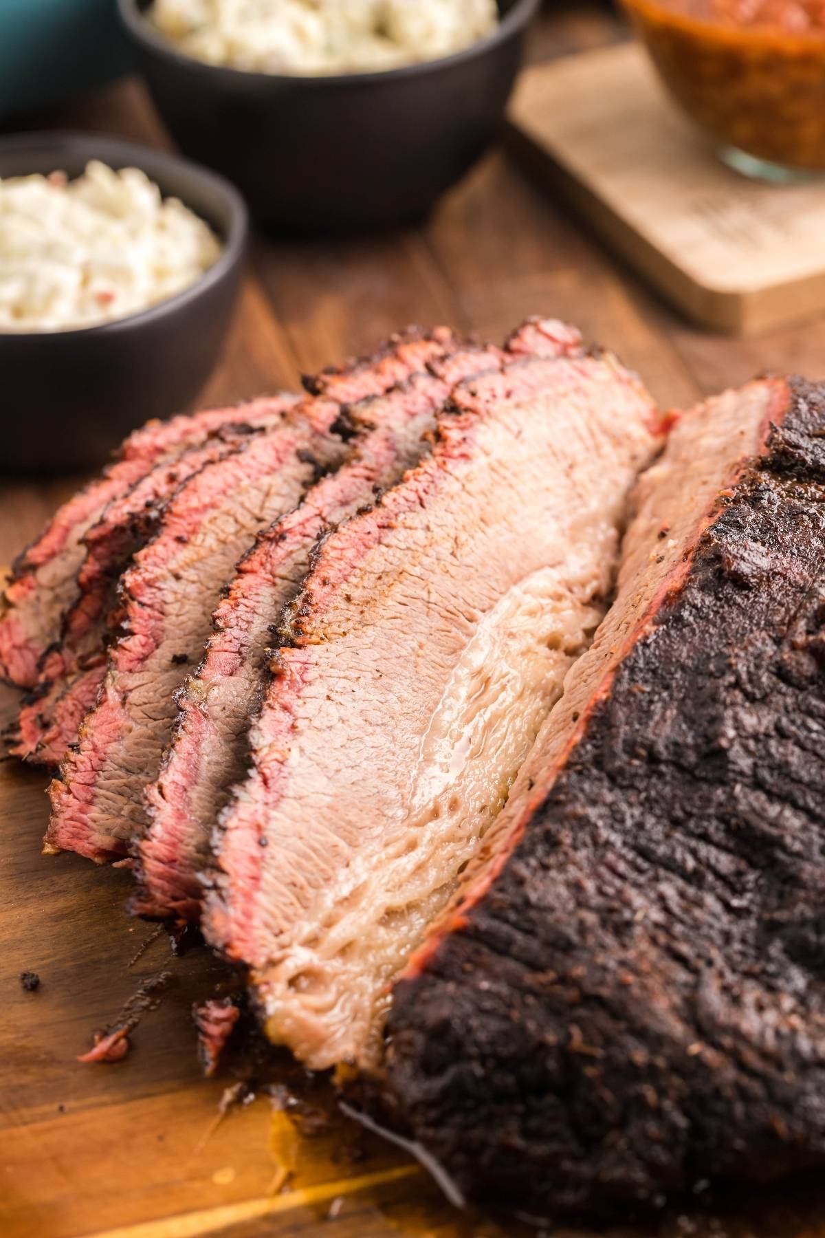 Mesquite smoked brisket, thinly sliced on a wooden board with bowls of side dishes in the background—perfect for anyone searching for an easy Texas smoked brisket recipe.