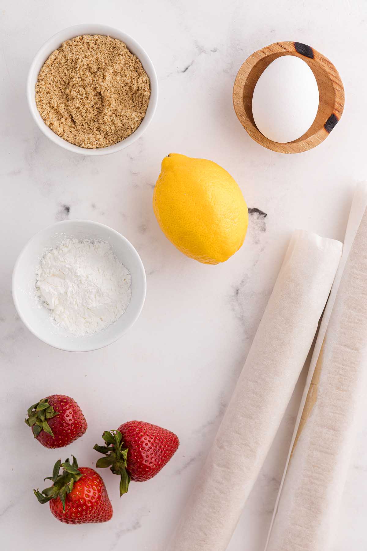 Bowl of brown sugar, egg, lemon, cornstarch, and strawberries on parchment paper—perfect for making mini strawberry pies on a white surface.